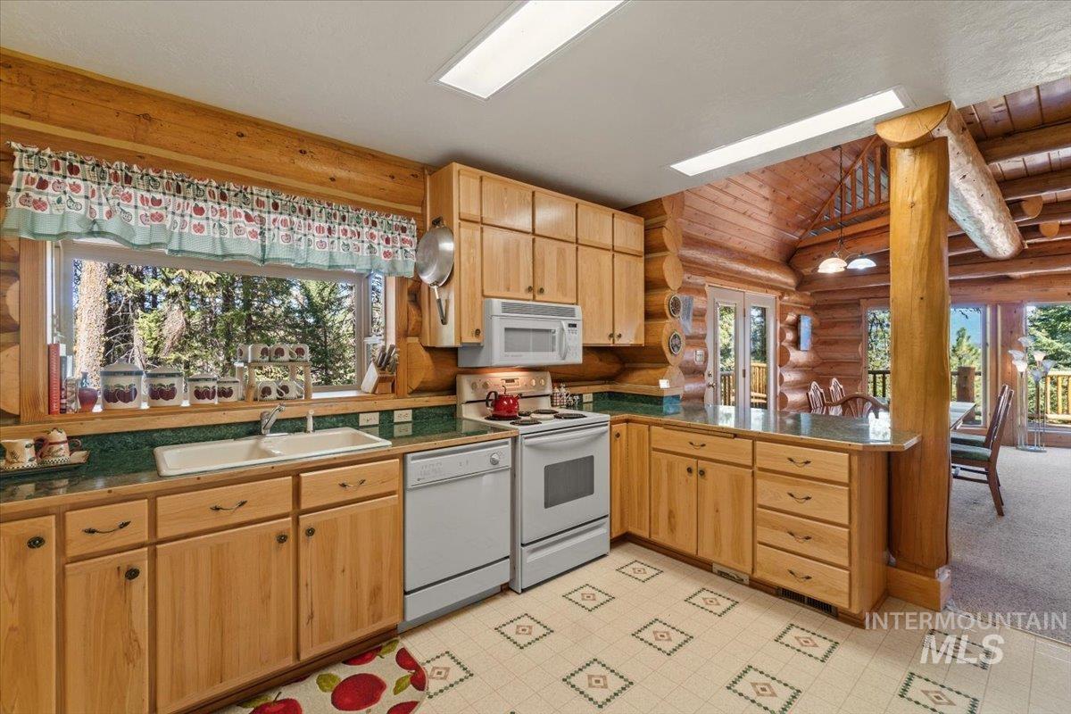 Kitchen with white appliances, log walls, light brown cabinetry, a peninsula, and dark countertops