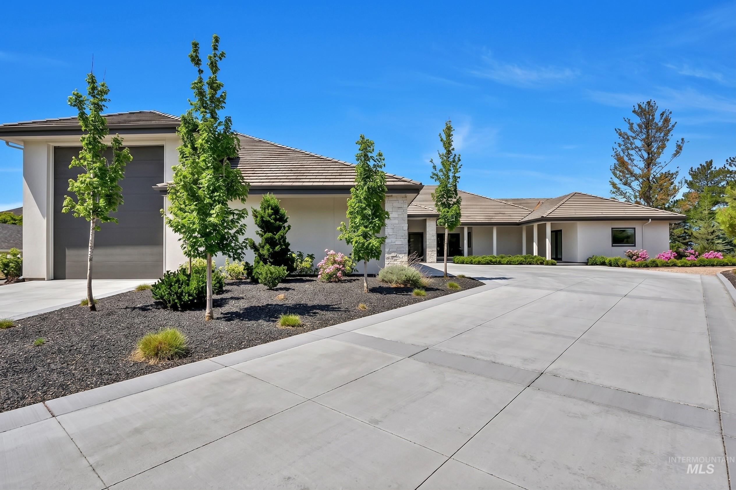 Contemporary stucco exterior with stone accents, a tile roof, an attached garage, a spacious concrete driveway, and established landscaping