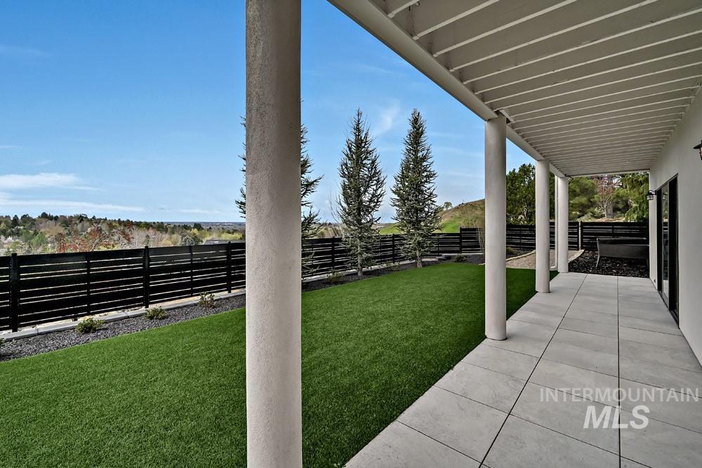 Covered patio featuring light-toned large format pavers, a manicured lawn, and a dark horizontal slat fence