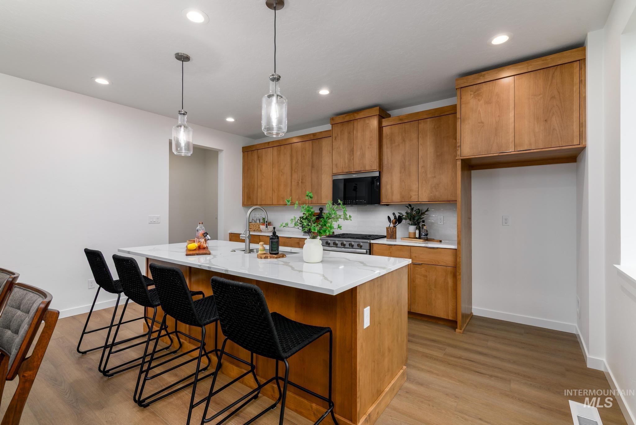 Kitchen featuring brown cabinetry, a center island with sink, pendant lighting, light stone counters, and a kitchen bar