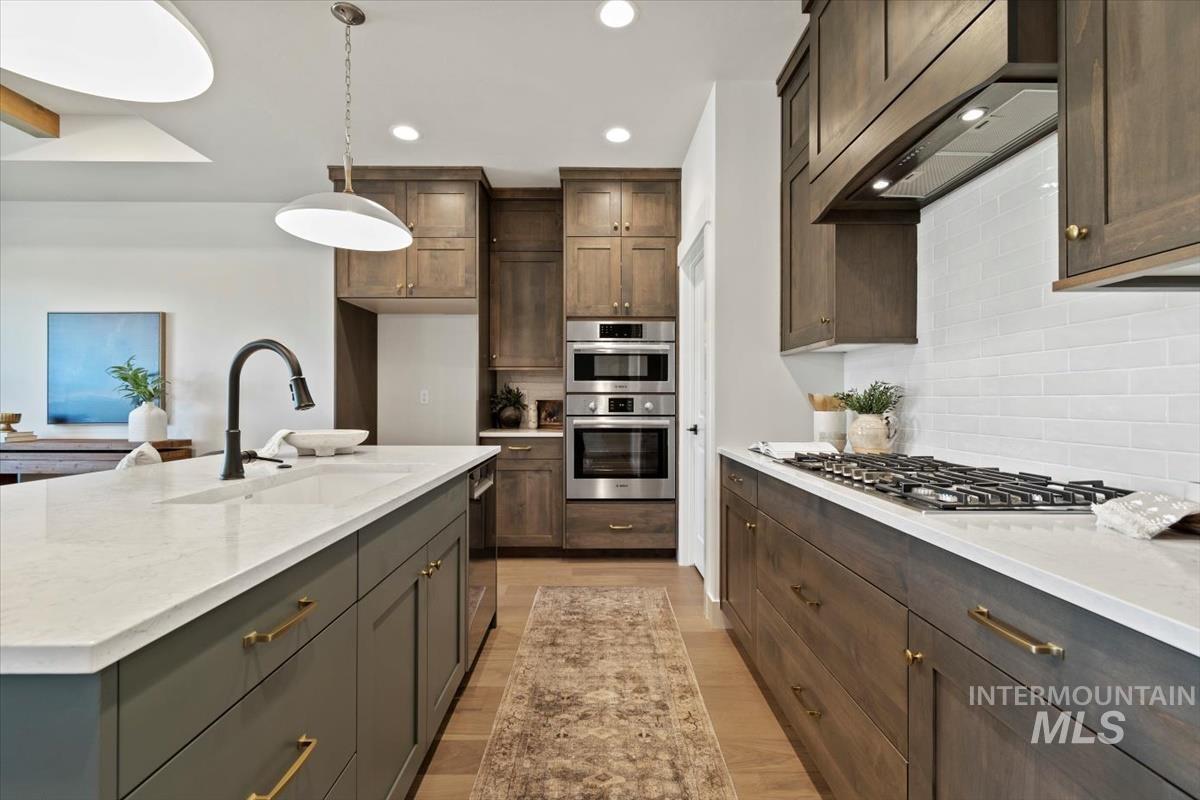 Kitchen featuring custom range hood, backsplash, light stone counters, appliances with stainless steel finishes, and light wood-style floors