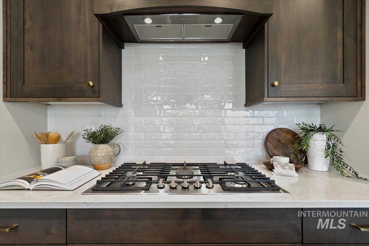 Kitchen featuring ventilation hood, dark brown cabinets, stainless steel gas cooktop, backsplash, and light stone countertops