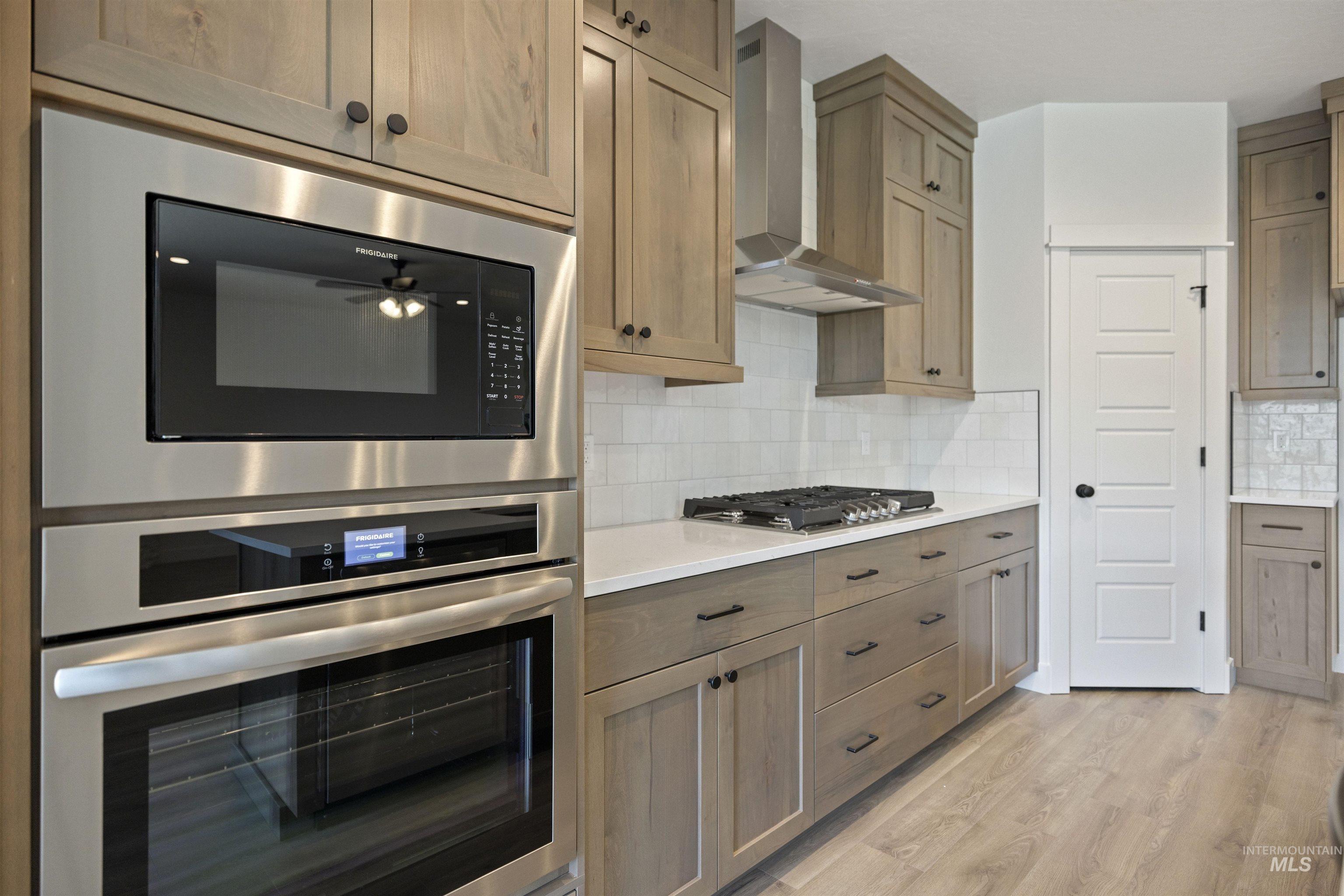 Kitchen featuring stainless steel appliances, wall chimney range hood, light wood-style flooring, light countertops, and decorative backsplash