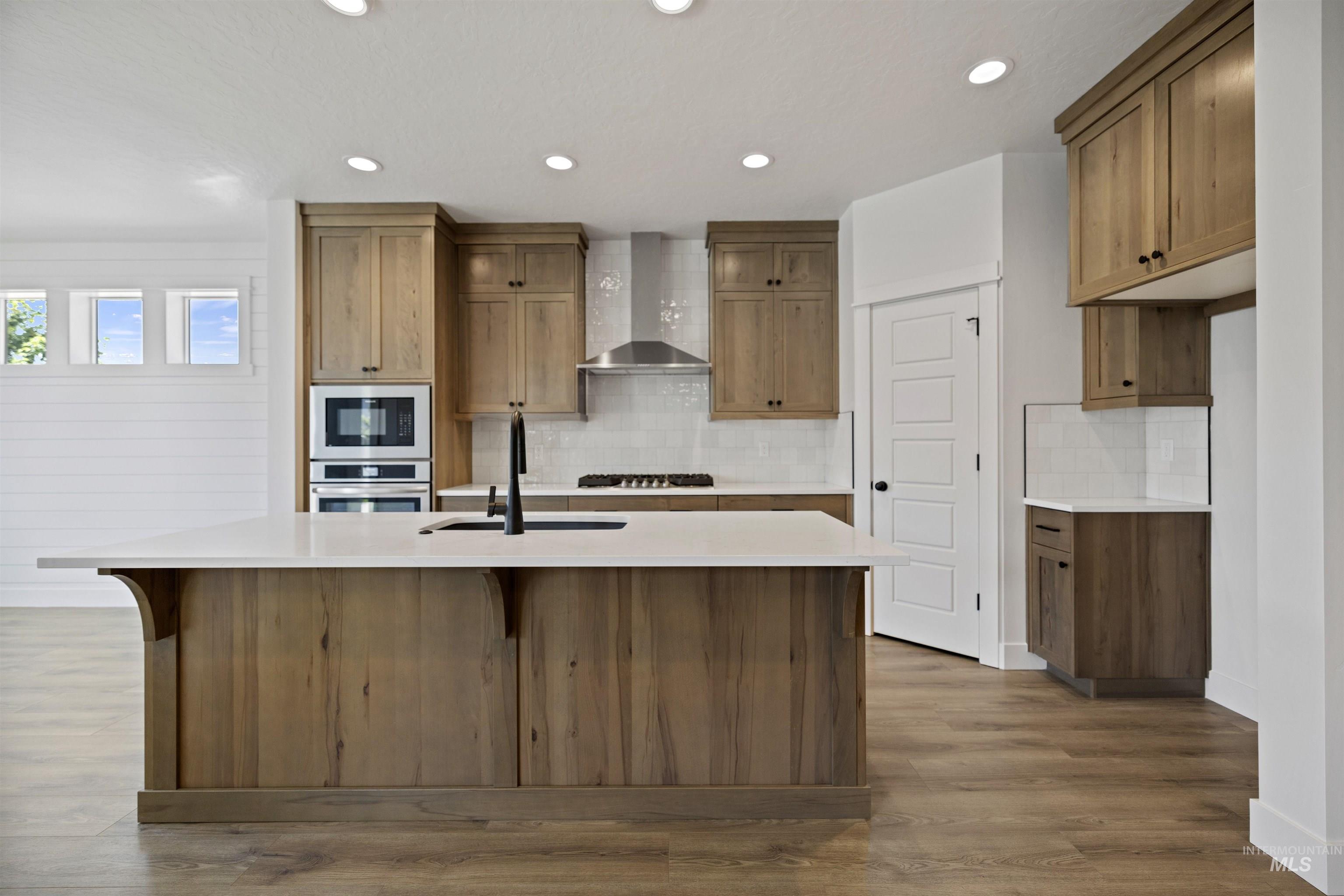 Kitchen with wall chimney range hood, an island with sink, decorative backsplash, light countertops, and stainless steel oven