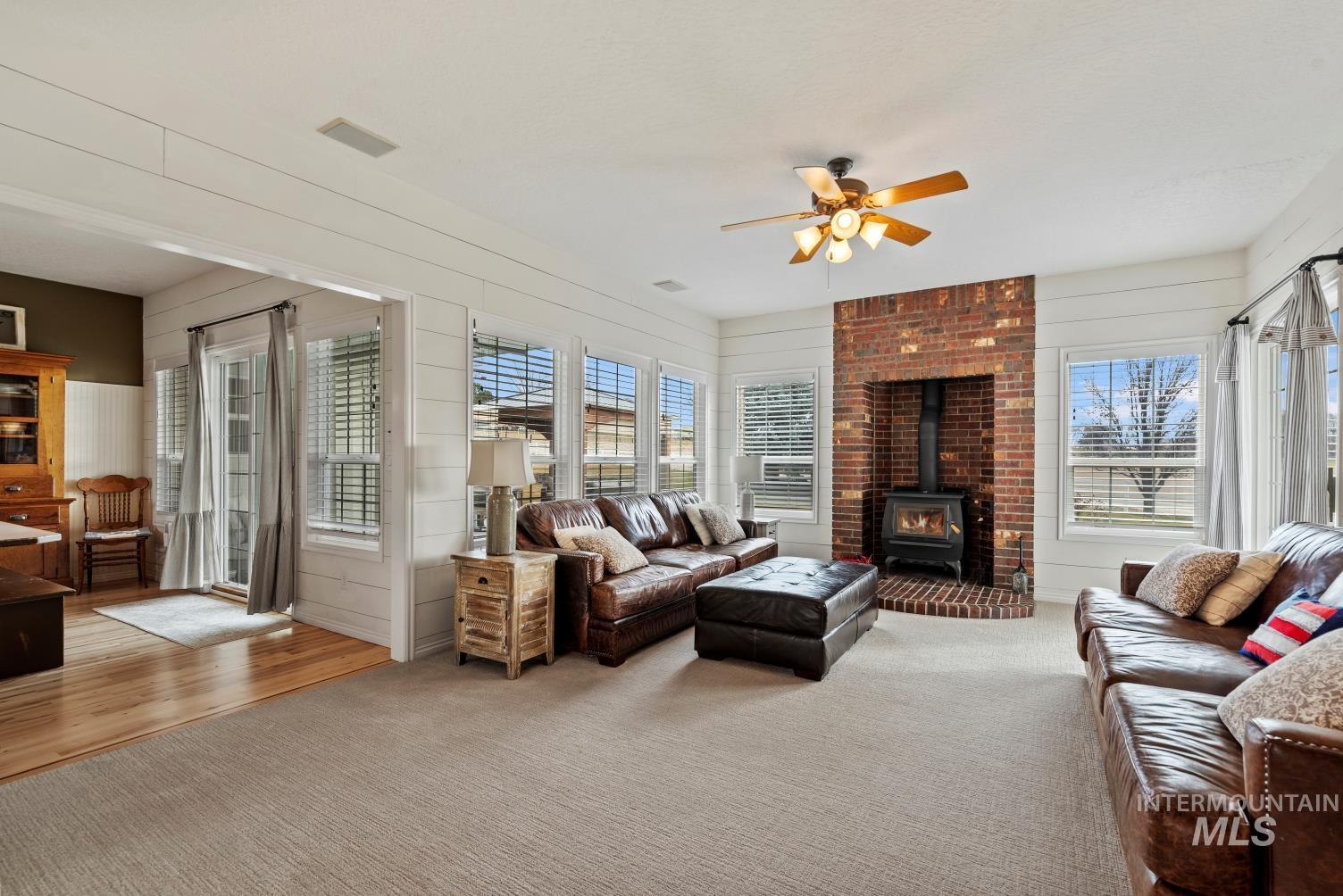 Living area with a ceiling fan, a wood stove, light colored carpet, and wooden walls