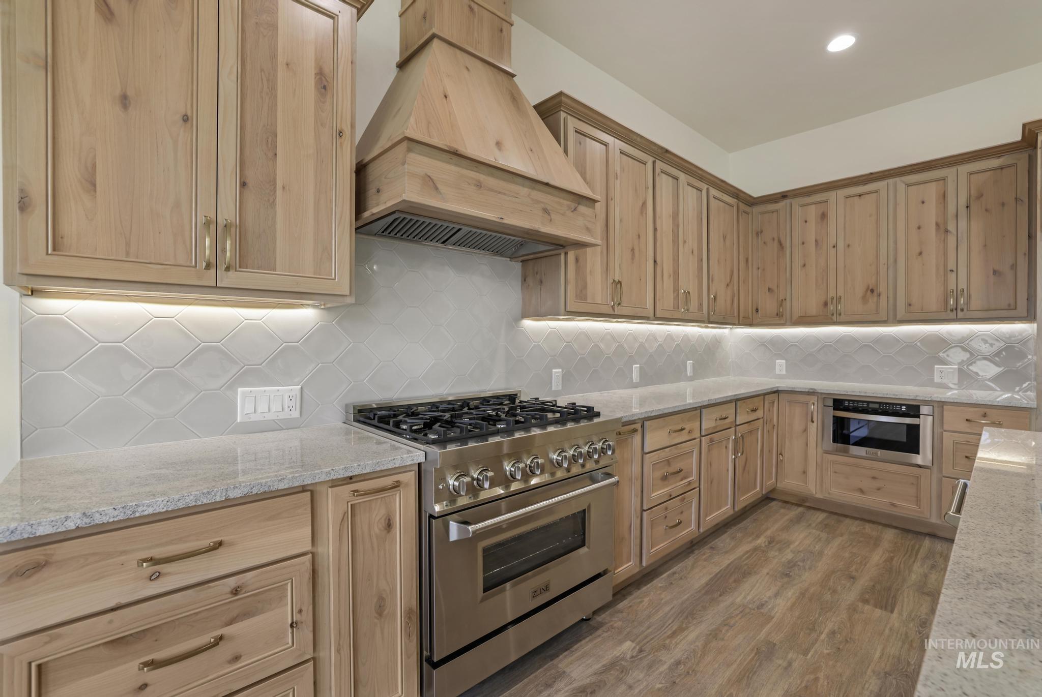 Kitchen with stainless steel appliances, custom exhaust hood, light stone counters, decorative backsplash, and light wood-style flooring