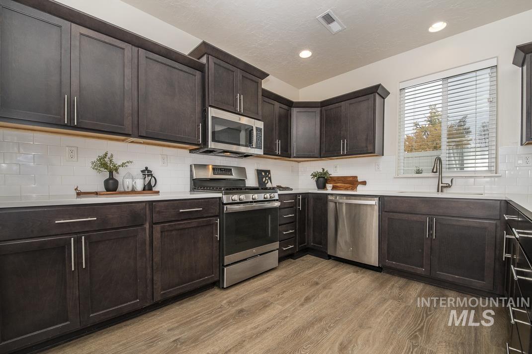 Kitchen featuring stainless steel appliances, dark brown cabinets, backsplash, light wood-type flooring, and light stone counters