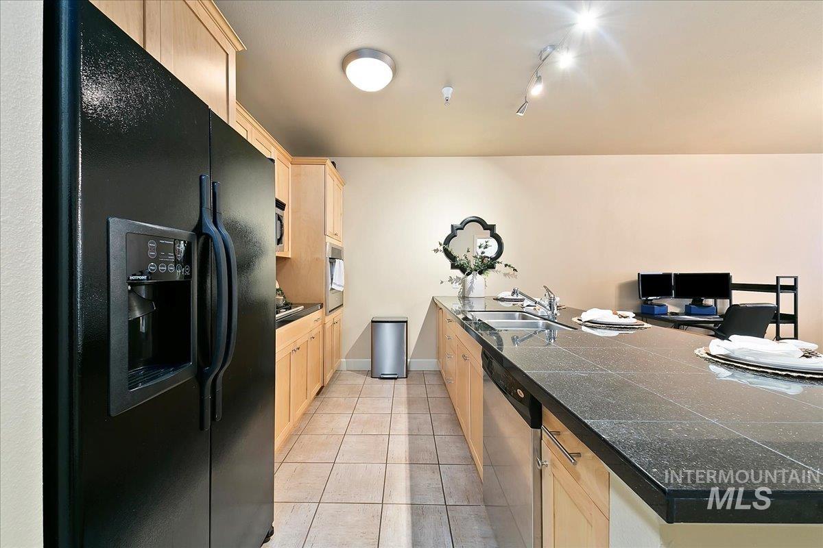 Kitchen with light wood finish cabinets, black fridge, stainless steel dishwasher, light tile patterned floors, and a peninsula