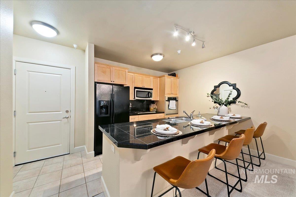 Kitchen with light wood finish cabinetry, stainless steel appliances, a breakfast bar area, tile counters, and light tile patterned floors