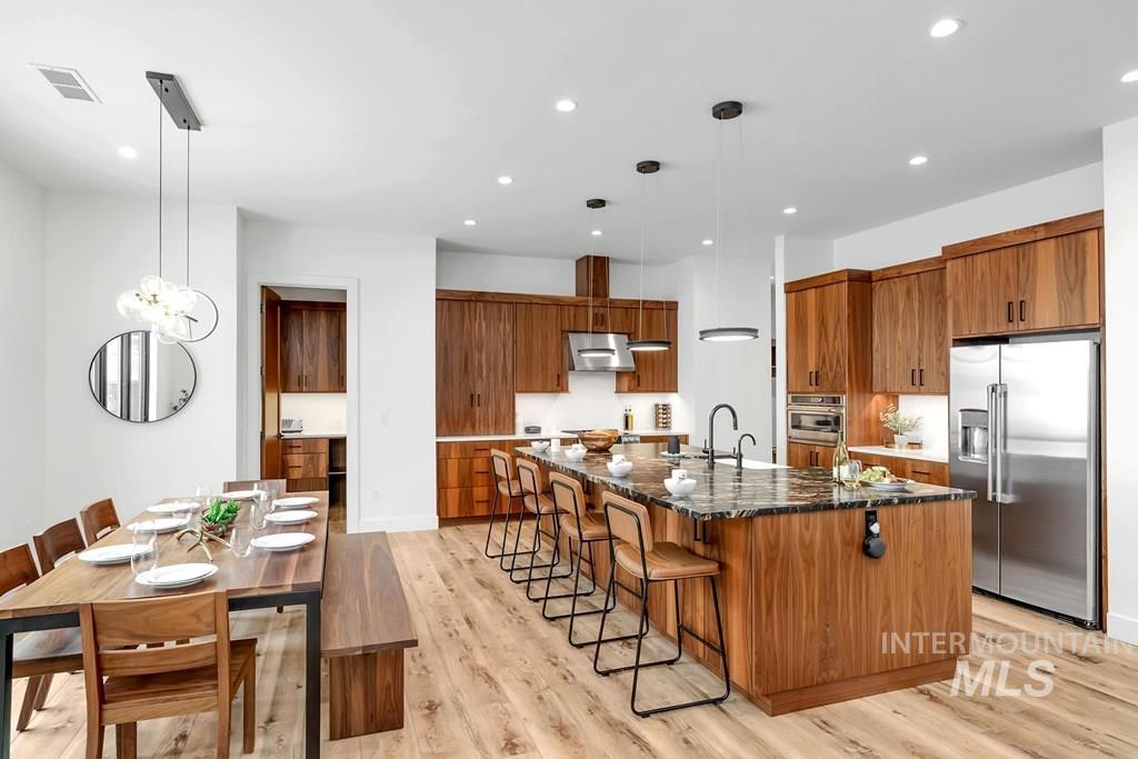 Kitchen featuring brown cabinets, appliances with stainless steel finishes, dark stone counters, light wood-type flooring, and hanging light fixtures