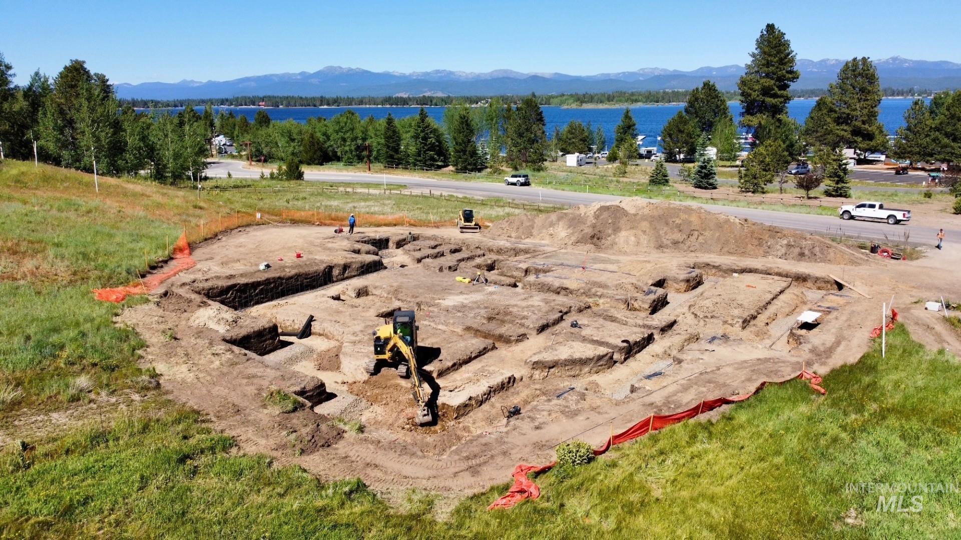Excavated building site featuring a prepared foundation, surrounding verdant grass, and a prominent water body with distant mountains