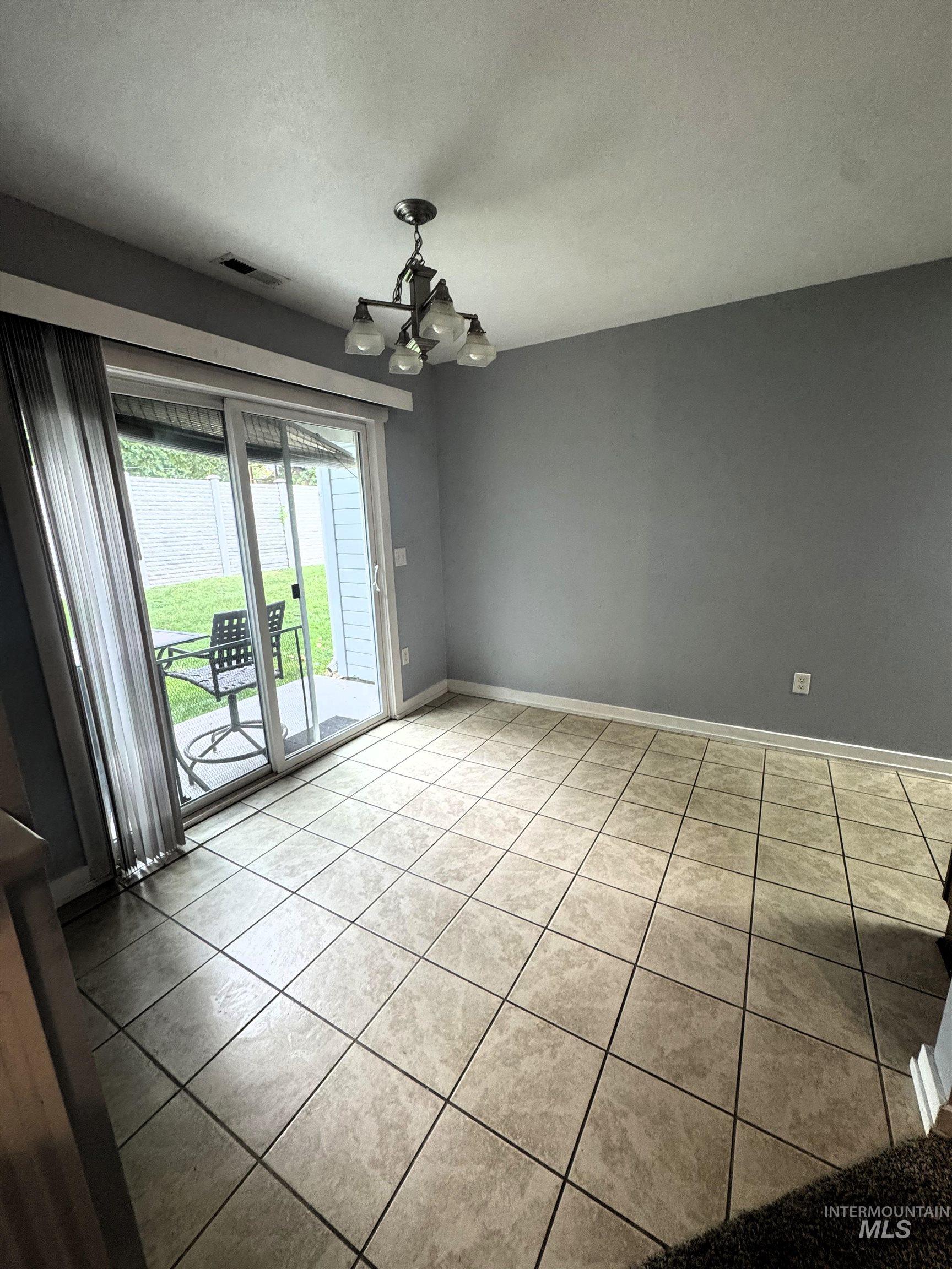 Spacious room featuring neutral-toned tile flooring, a contemporary chandelier, and a sliding glass door providing access to an outdoor patio area
