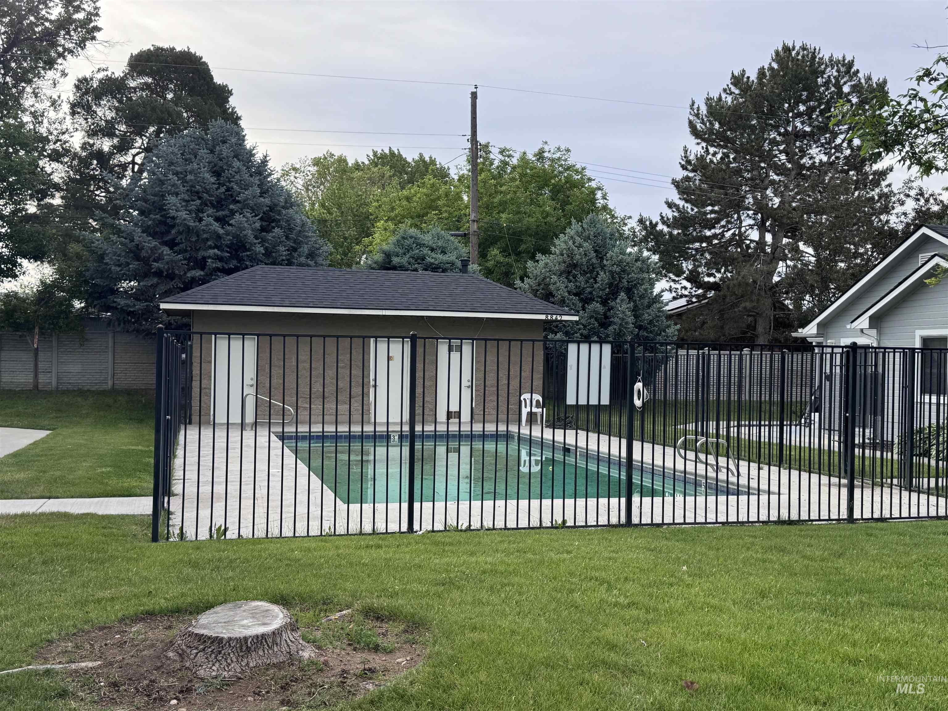 Fenced swimming pool area with concrete decking and adjacent pool house featuring a dark shingle roof and two entry doors