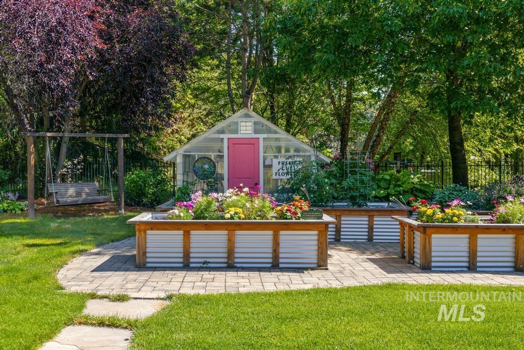 Landscaped garden featuring a clear-paneled greenhouse with a pink door, multiple raised garden beds with corrugated metal and wood trim, and a stone paver patio