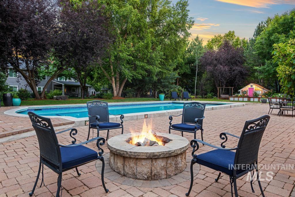 Expansive paver patio featuring a built-in stone fire pit, a large inground swimming pool, and mature trees providing natural screening