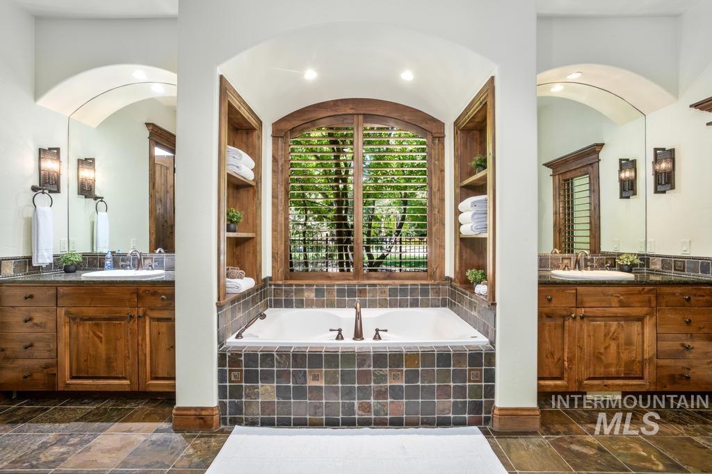 Soaking tub with tile surround, flanked by built-in wood shelving and an arched window with plantation shutters