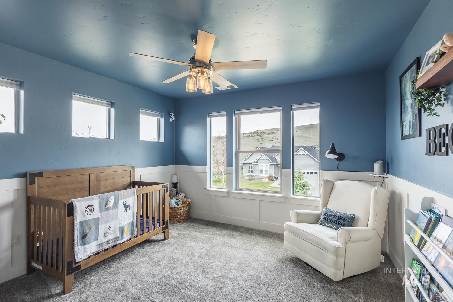 Carpeted bedroom with a crib, a wainscoted wall, a ceiling fan, and a decorative wall