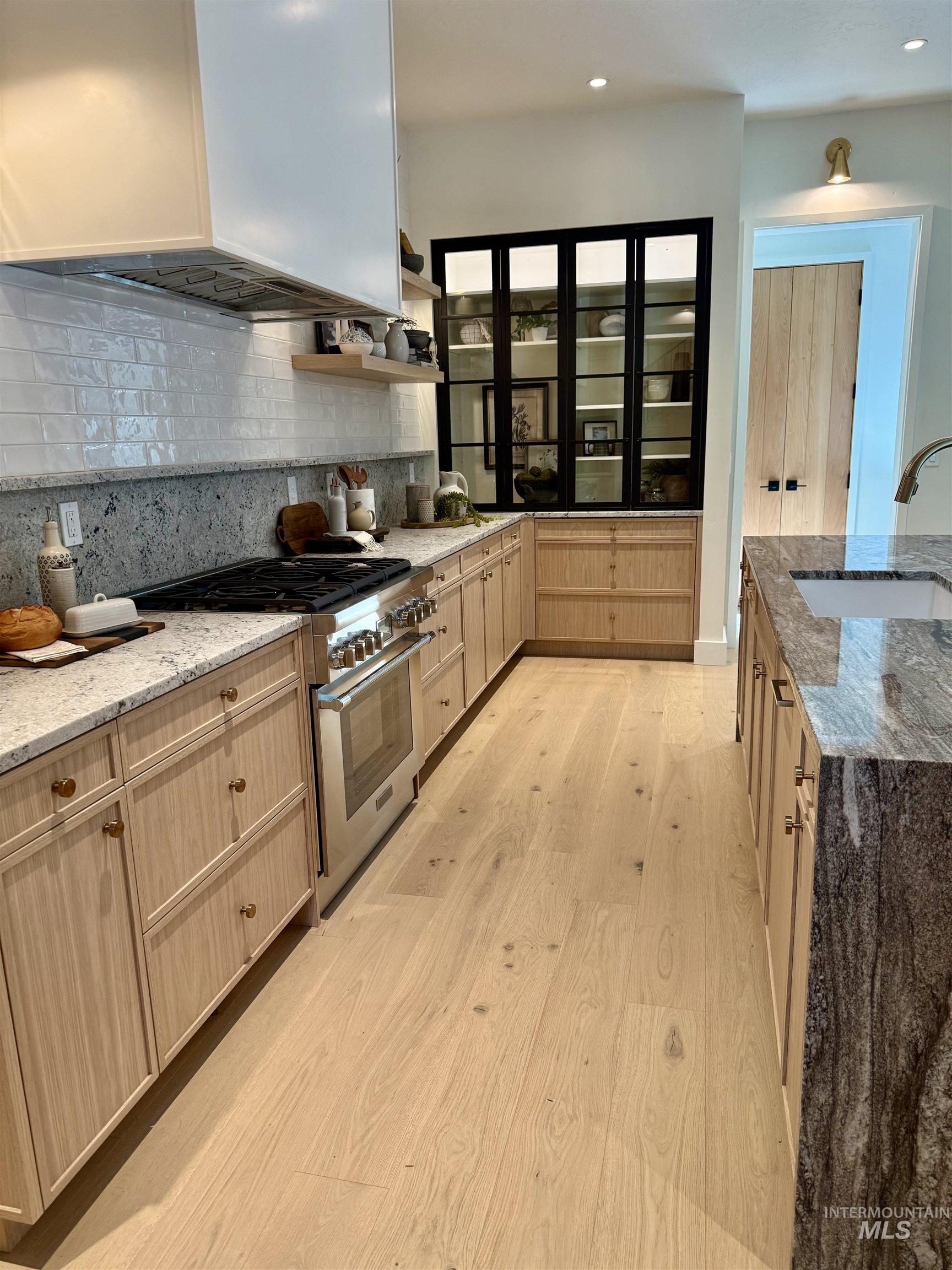 Kitchen with light stone countertops, light brown cabinets, stainless steel stove, tasteful backsplash, and wall chimney exhaust hood