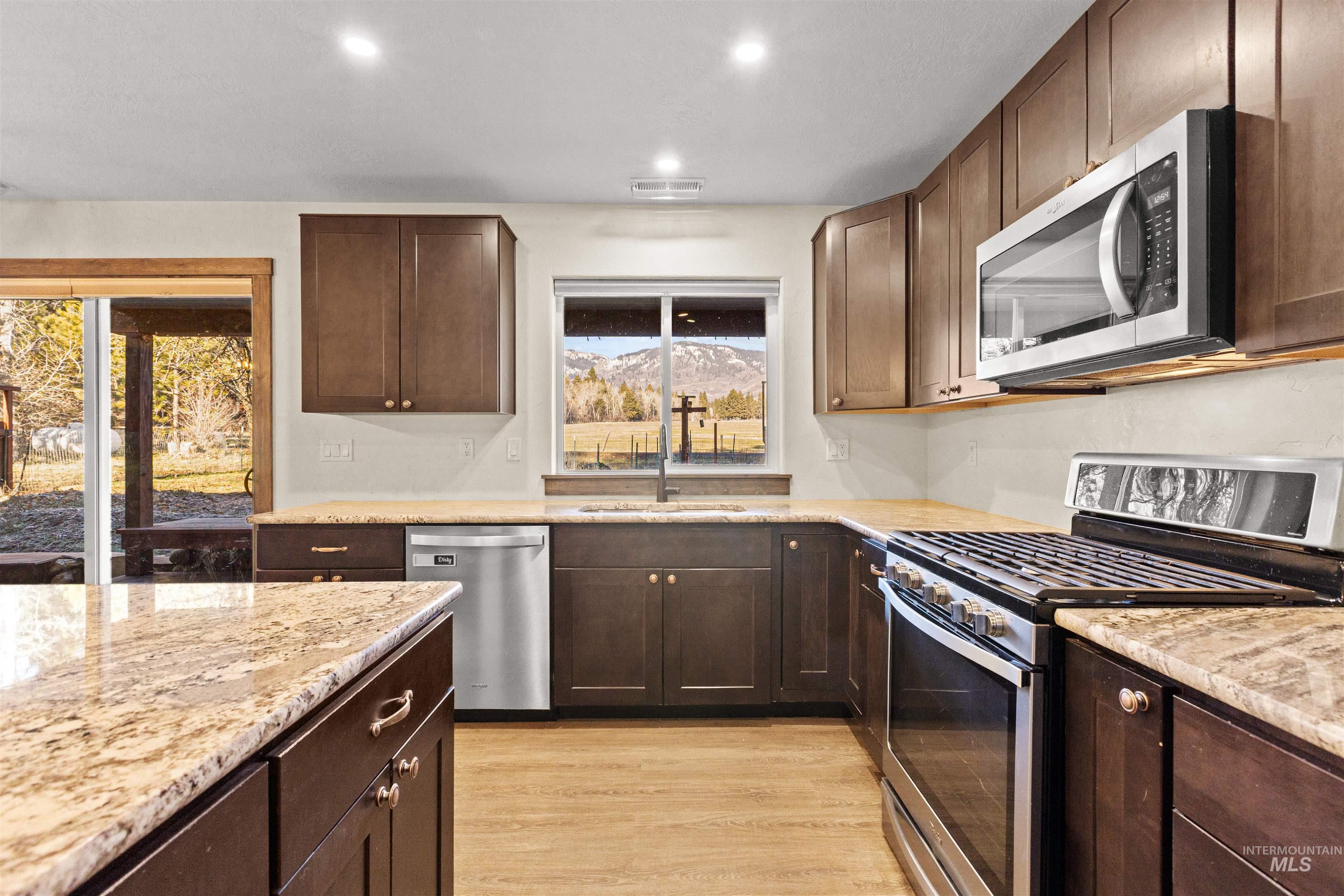Kitchen with dark brown cabinetry, stainless steel appliances, light stone counters, light wood-style flooring, and recessed lighting