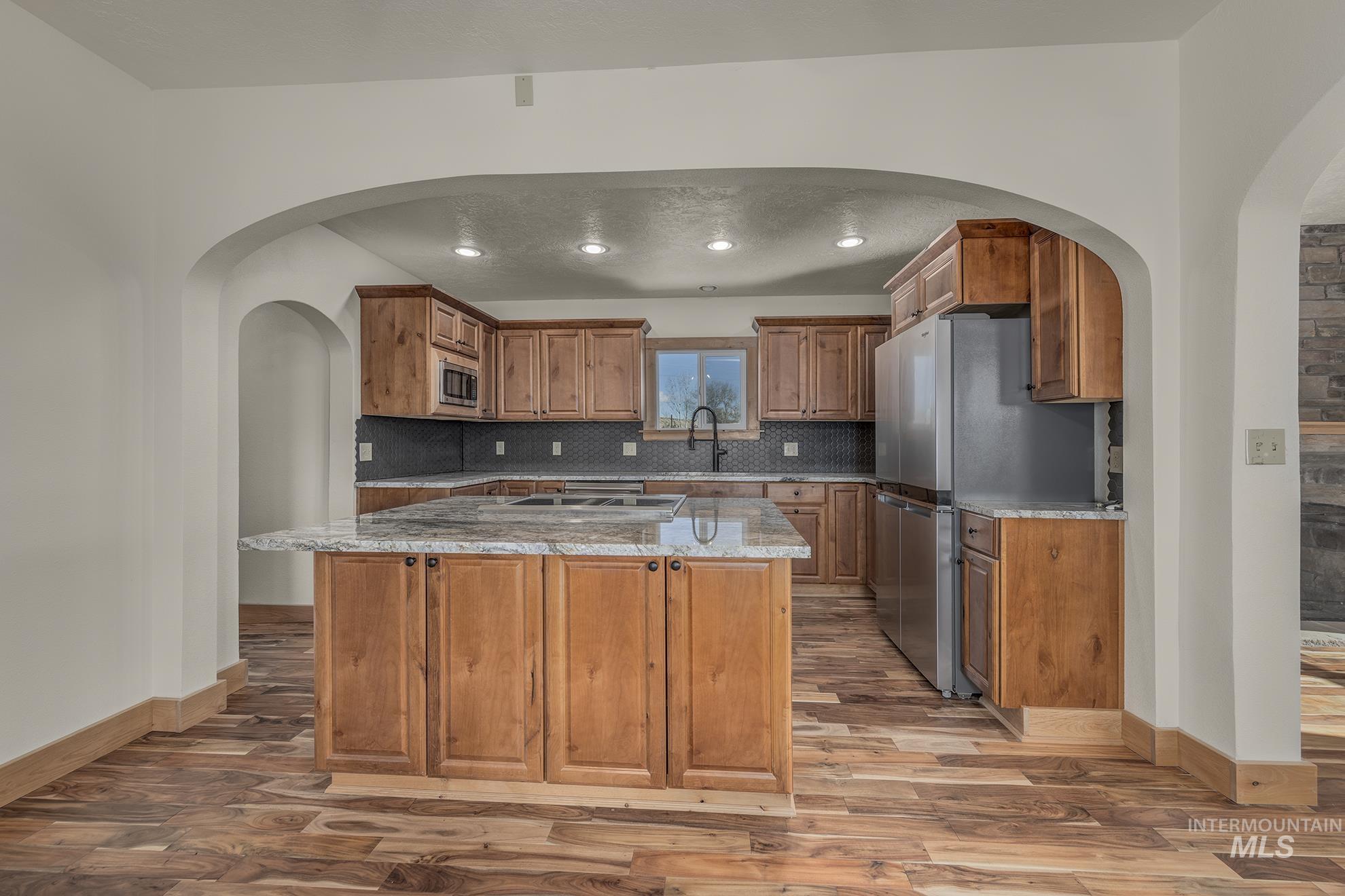 Kitchen with arched walkways, brown cabinetry, light stone countertops, stainless steel appliances, and a center island