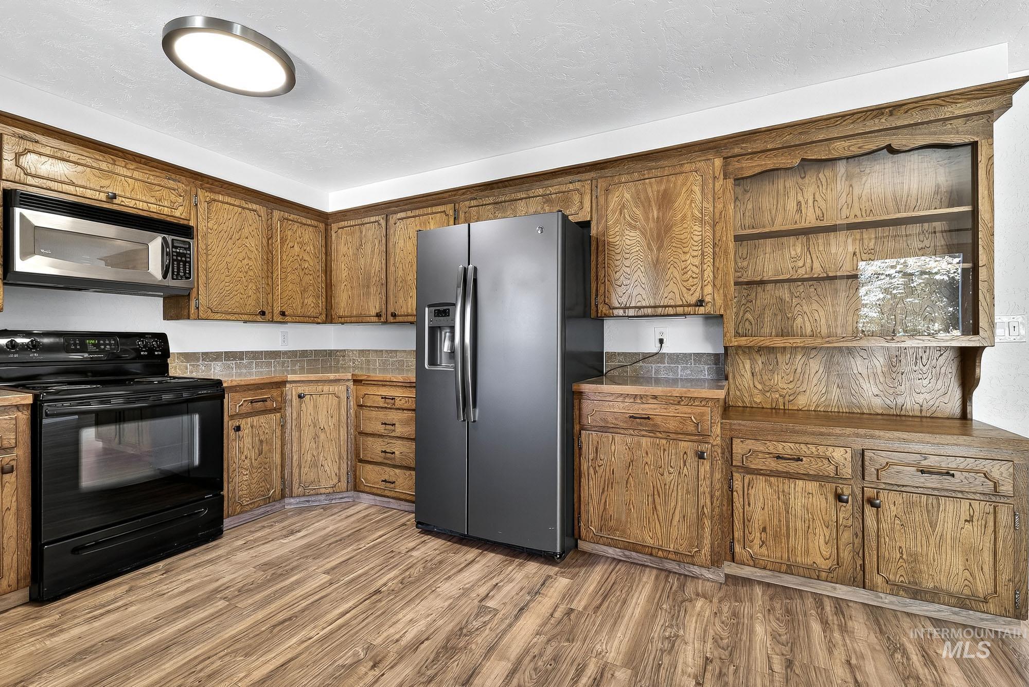 Kitchen with open shelves, appliances with stainless steel finishes, brown cabinets, light wood-style flooring, and a textured ceiling