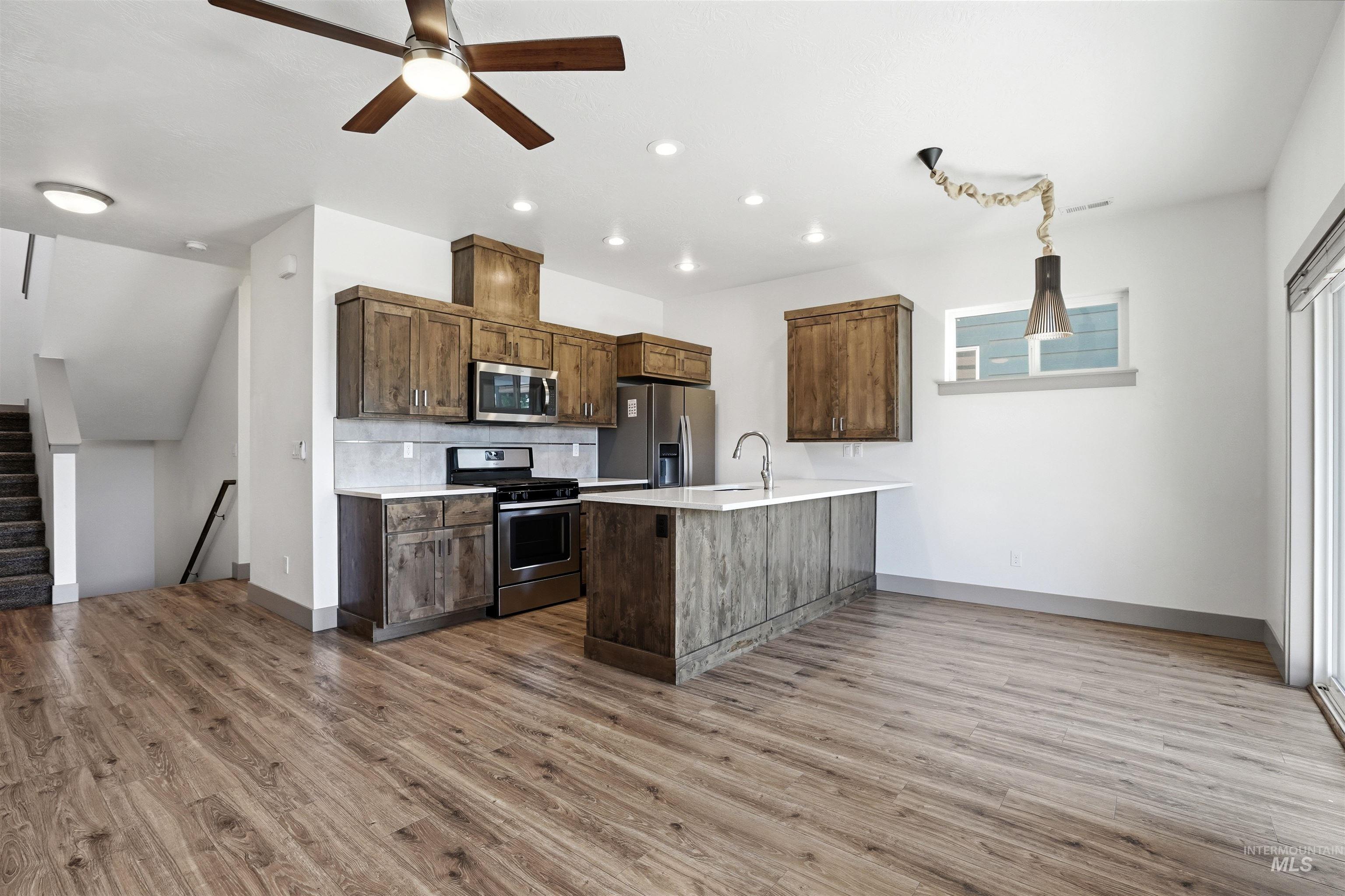 Modern kitchen featuring wood-finish cabinetry, stainless steel appliances, a white countertop island with an integrated sink, recessed lighting, and wood-finish flooring