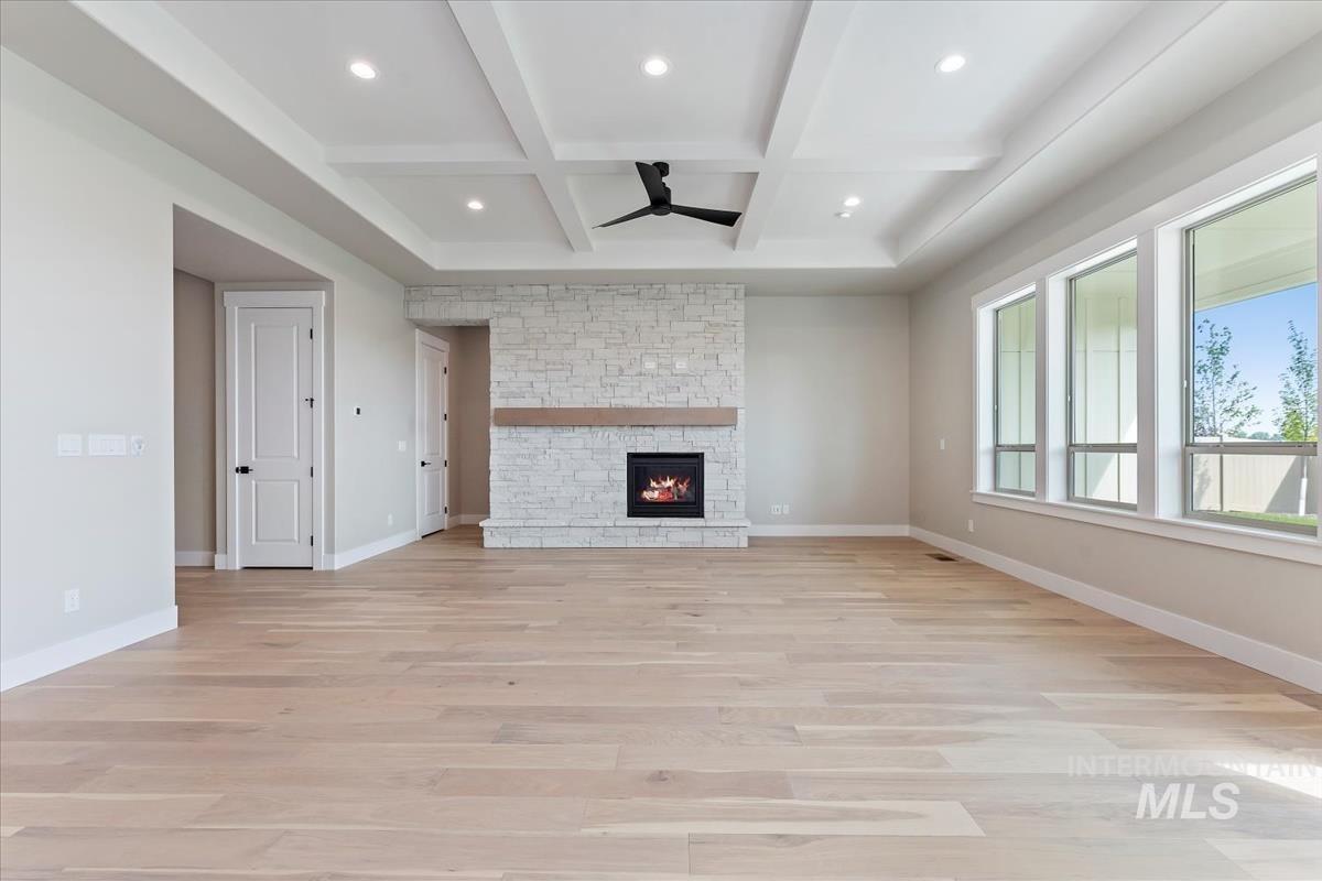Unfurnished living room featuring beam ceiling, light wood-style floors, a stone fireplace, coffered ceiling, and a ceiling fan