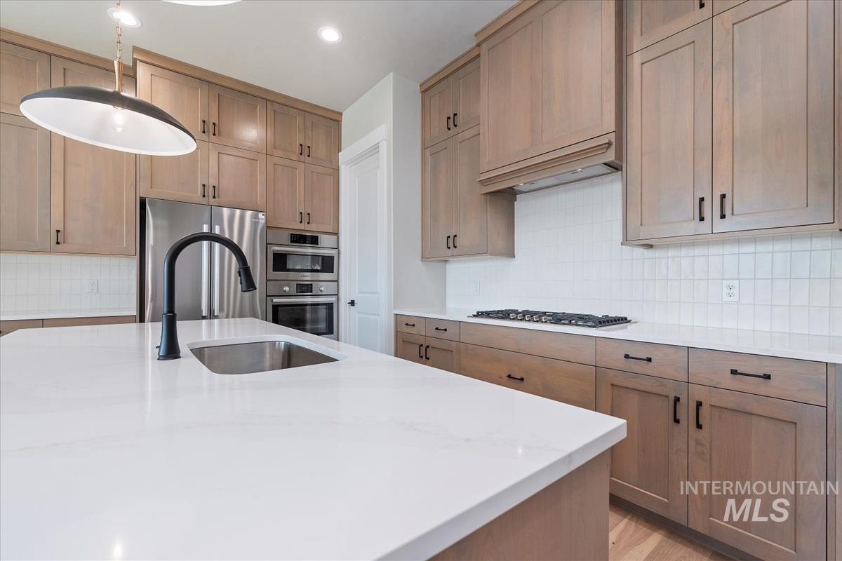 Kitchen with backsplash, light stone countertops, appliances with stainless steel finishes, decorative light fixtures, and brown cabinetry