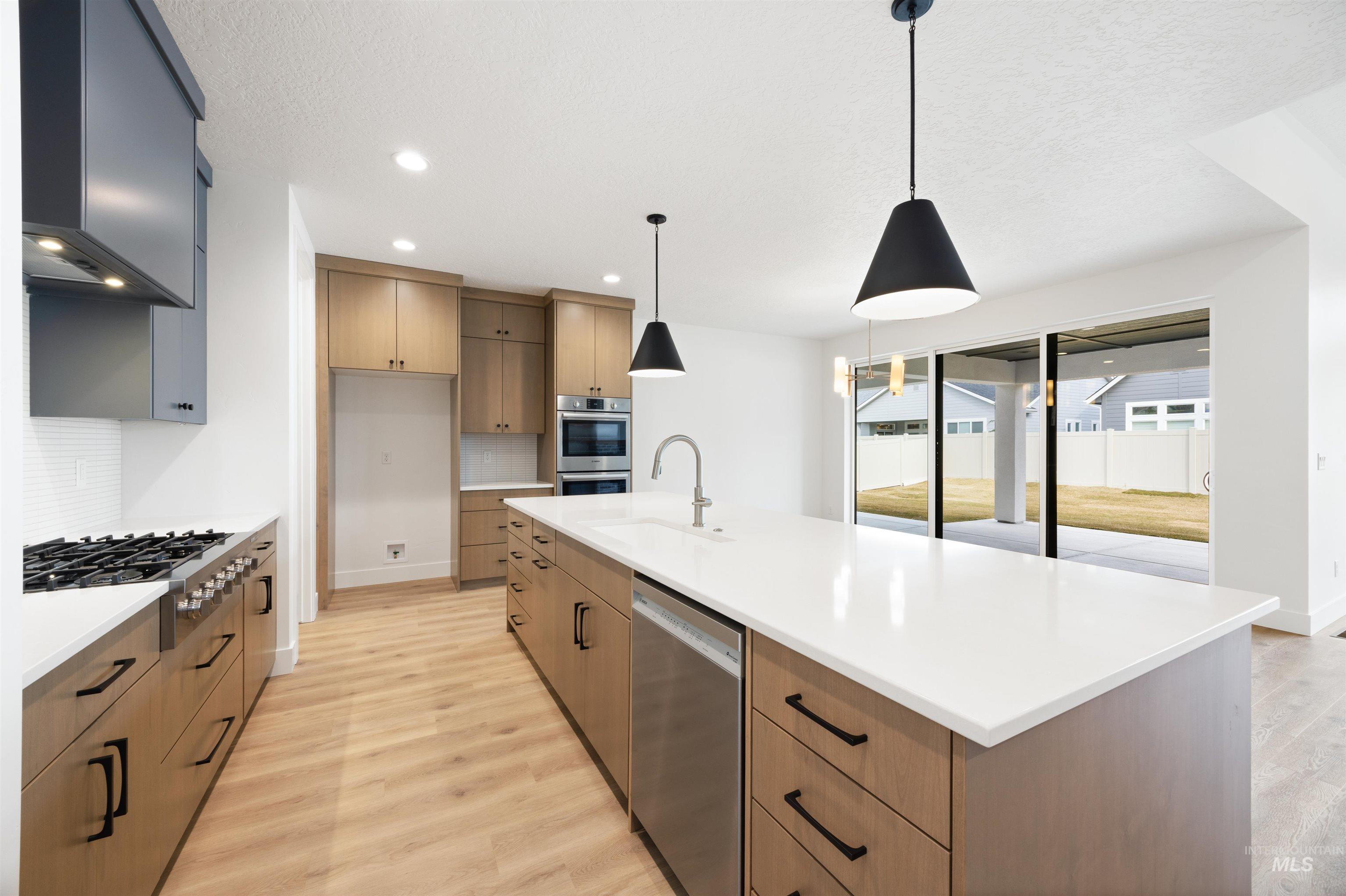 Kitchen with light wood-style flooring, hanging light fixtures, stainless steel appliances, ventilation hood, and an island with sink