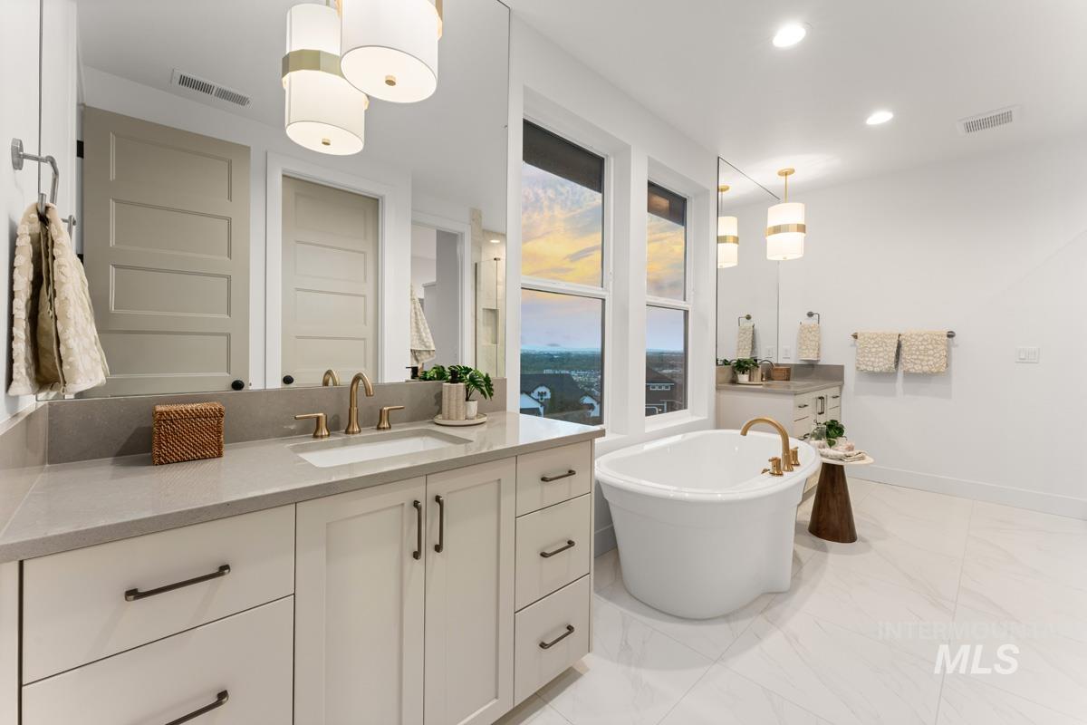 Bright bathroom featuring a freestanding tub with brass fixtures, dual vanities with quartz countertops, custom cabinetry, and large windows