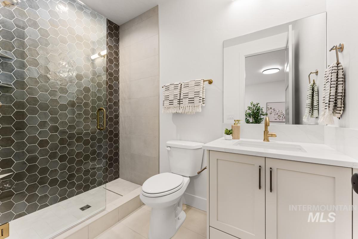 Shower with hexagonal tile surround and glass enclosure, white vanity with quartz countertop, undermount sink, and brass-finish faucet