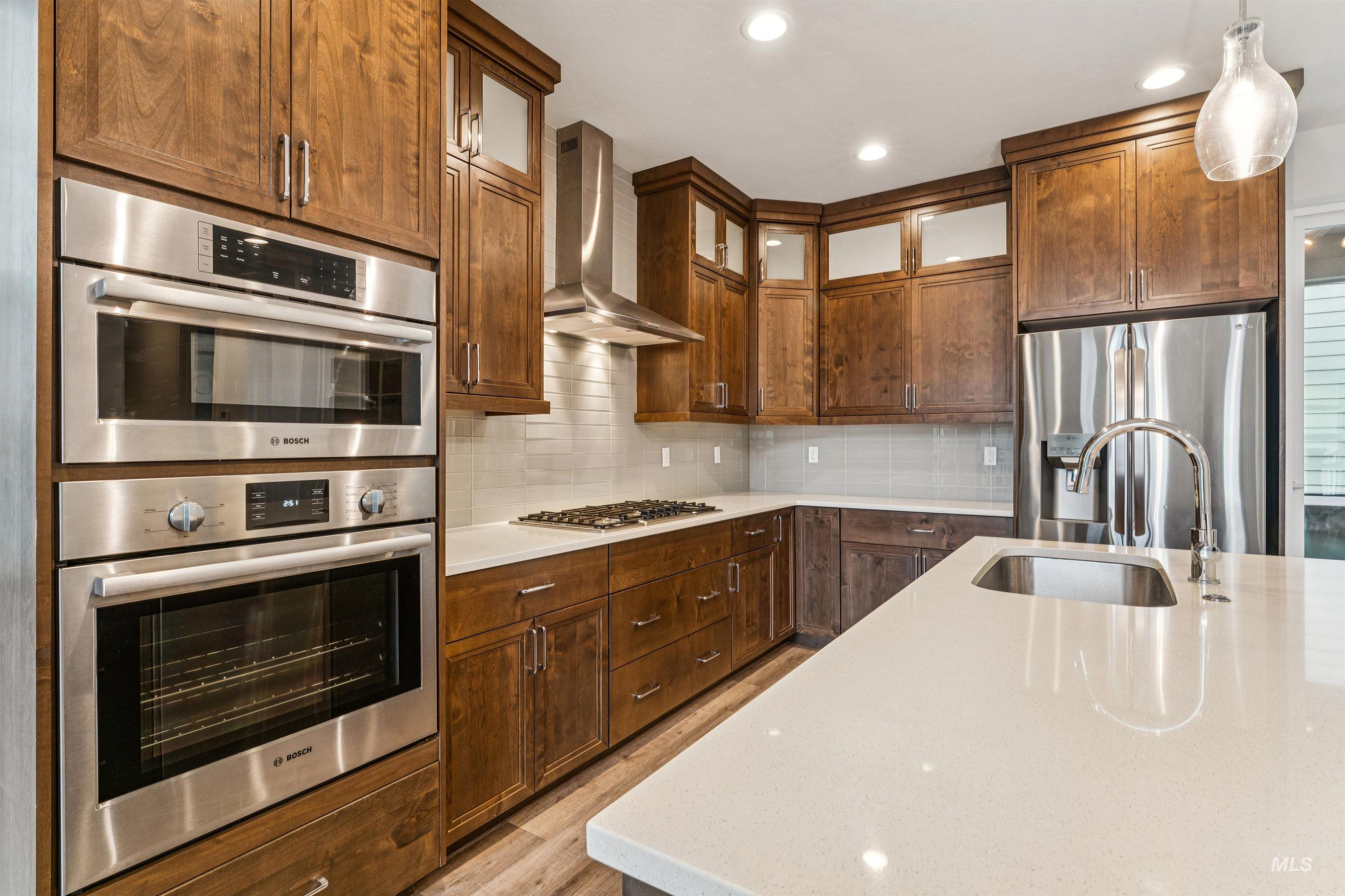 Kitchen with hanging light fixtures, tasteful backsplash, wall chimney exhaust hood, appliances with stainless steel finishes, and light quartz countertops