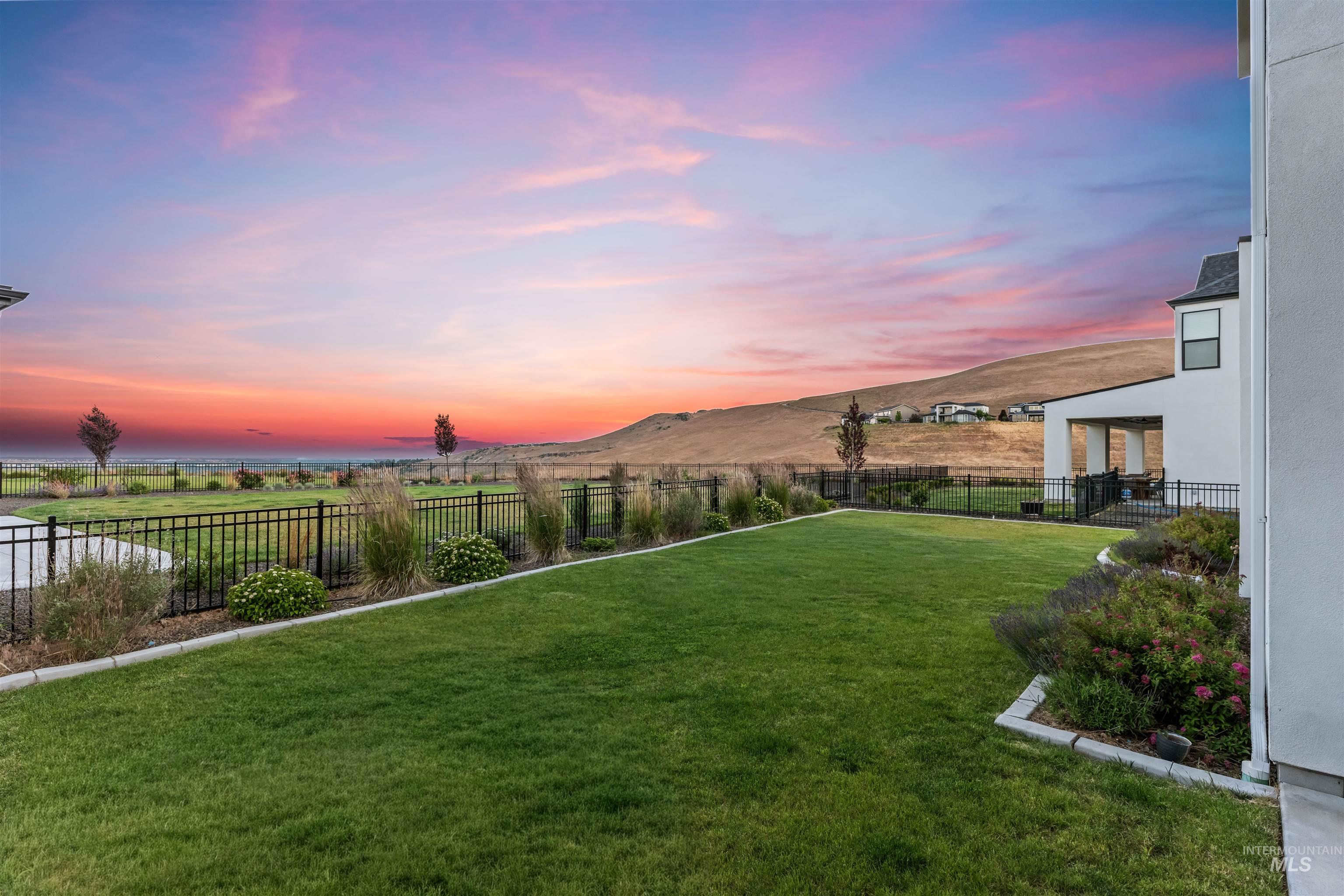 Expansive grassy backyard featuring a black metal fence, landscaped planting beds, and a concrete patio