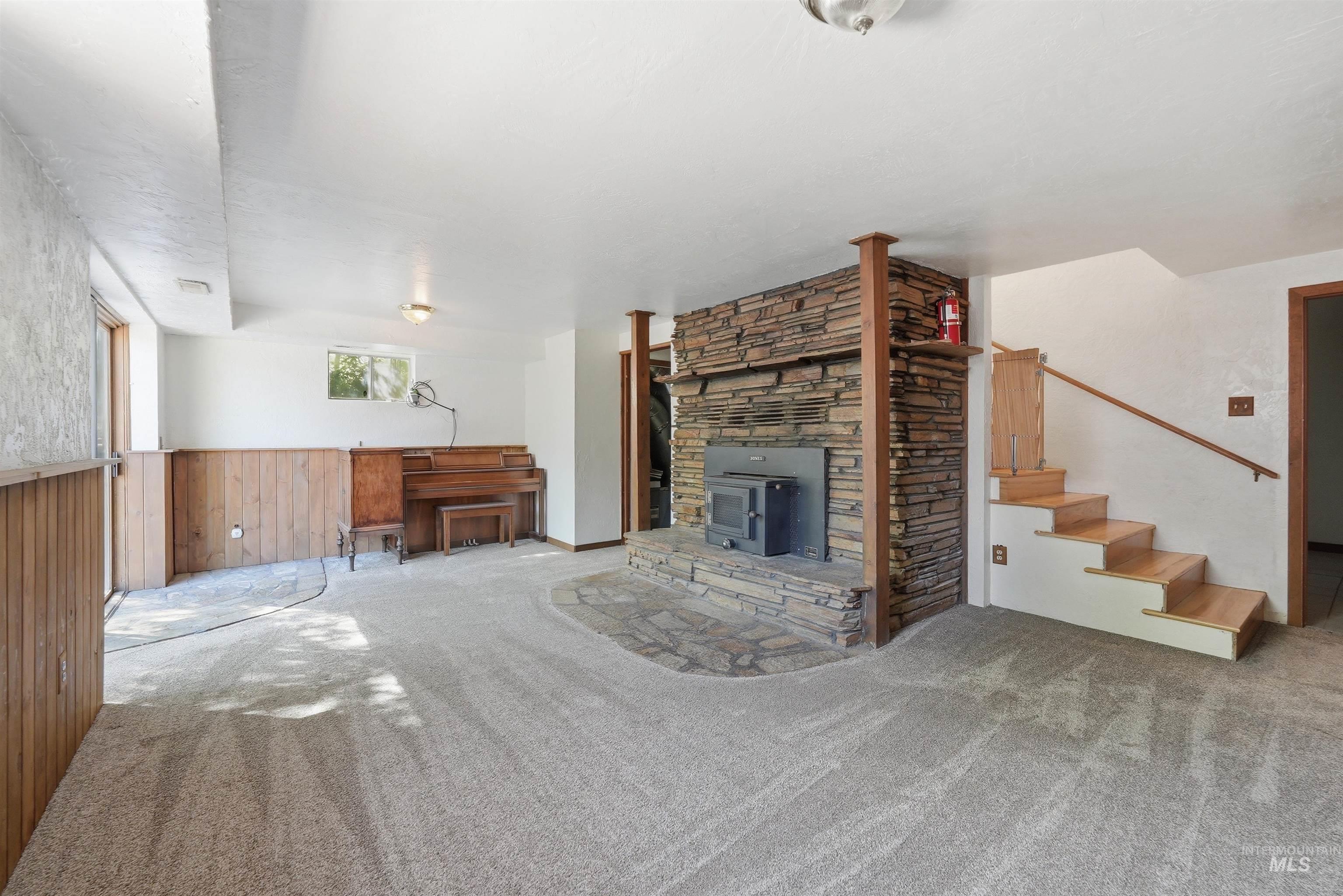 Spacious room featuring a stone-clad fireplace with wood-burning insert, wood-paneled wainscoting, light-toned carpet, a wooden staircase, and natural light from a window