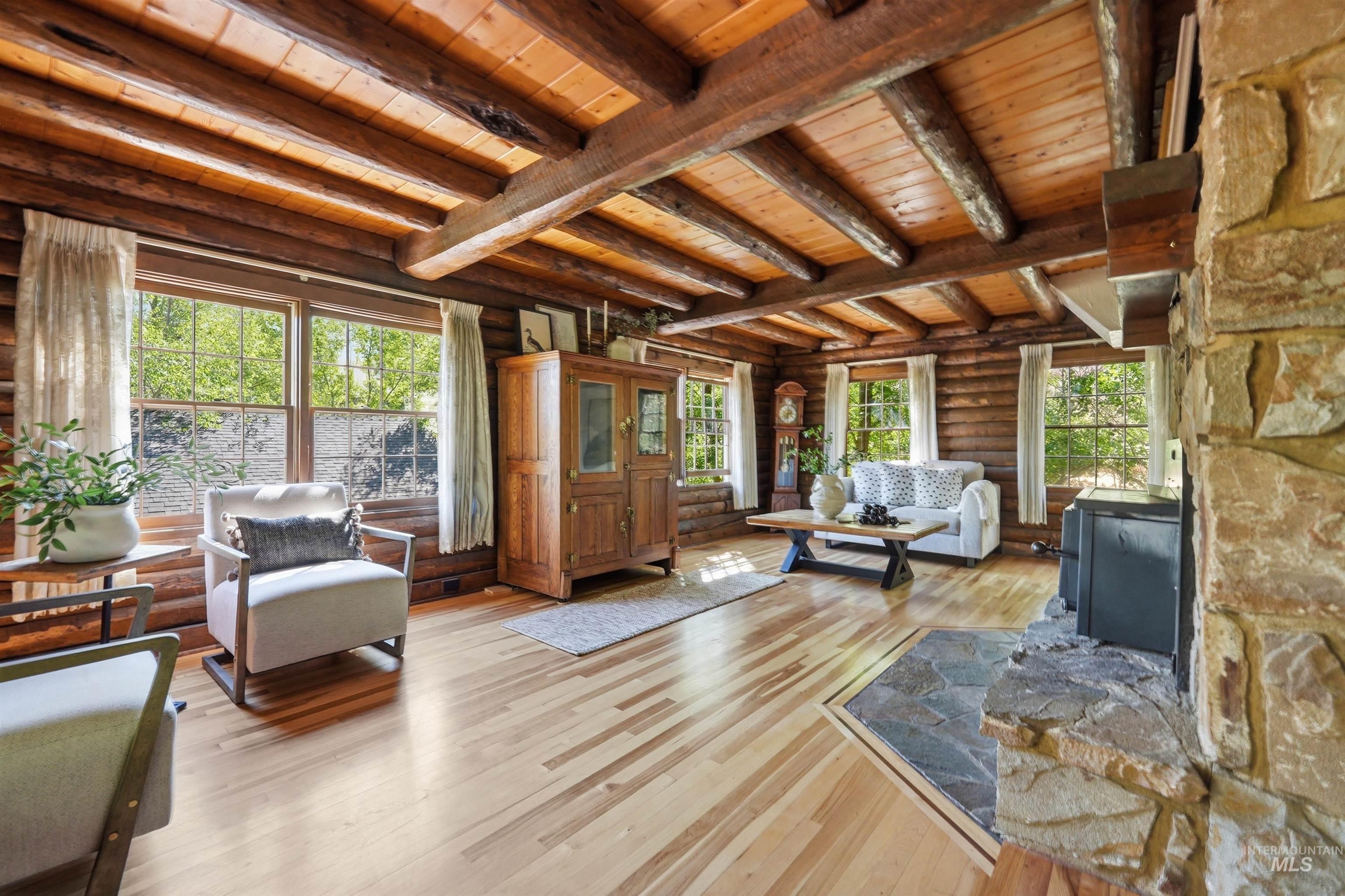 Living area featuring exposed wooden beams, natural log walls, light wood-finish flooring, a stone fireplace with hearth, and multiple windows