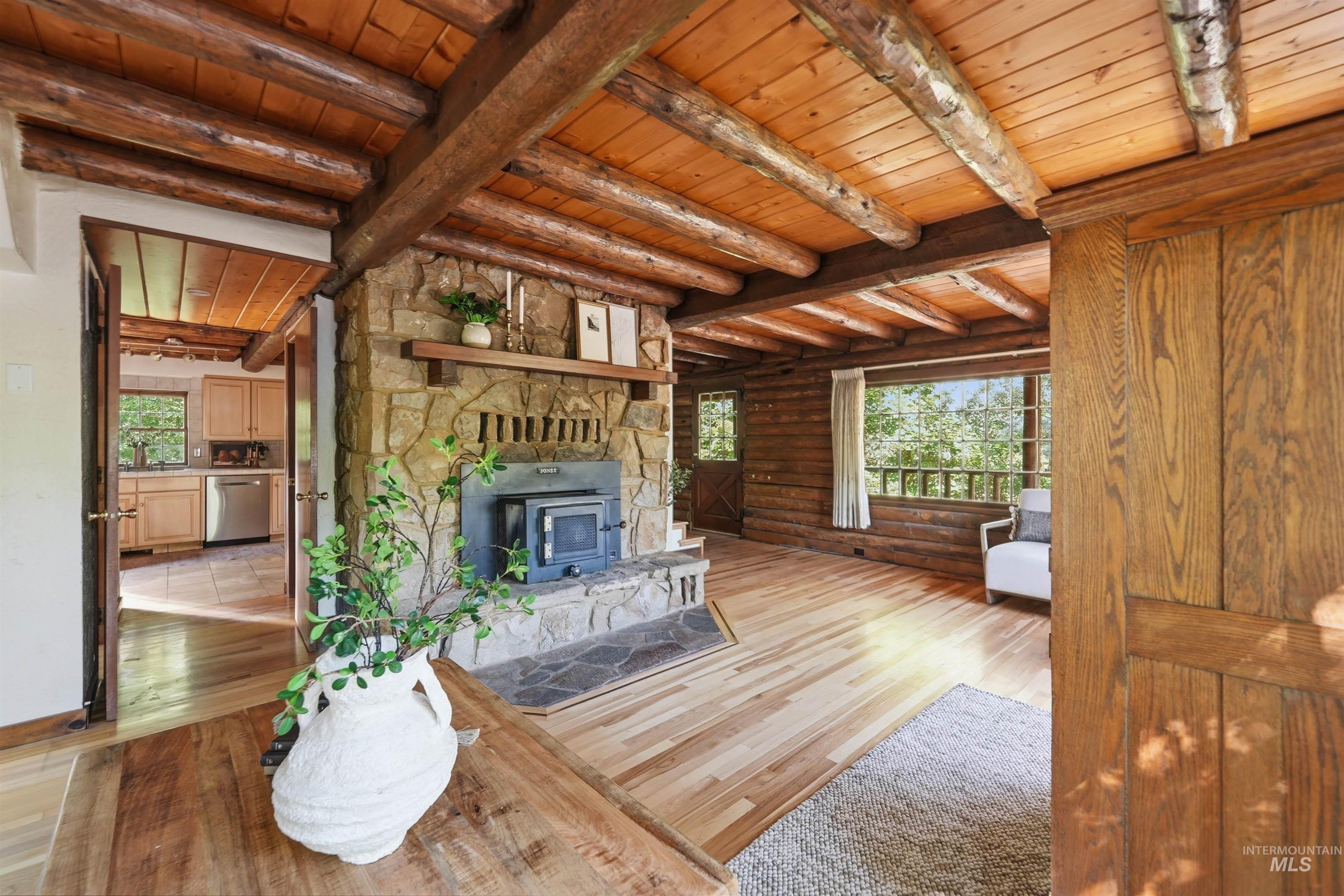 Spacious living area featuring a stone-clad fireplace with wood-burning insert, exposed log beam ceiling, light wood-finish flooring, and a stone hearth