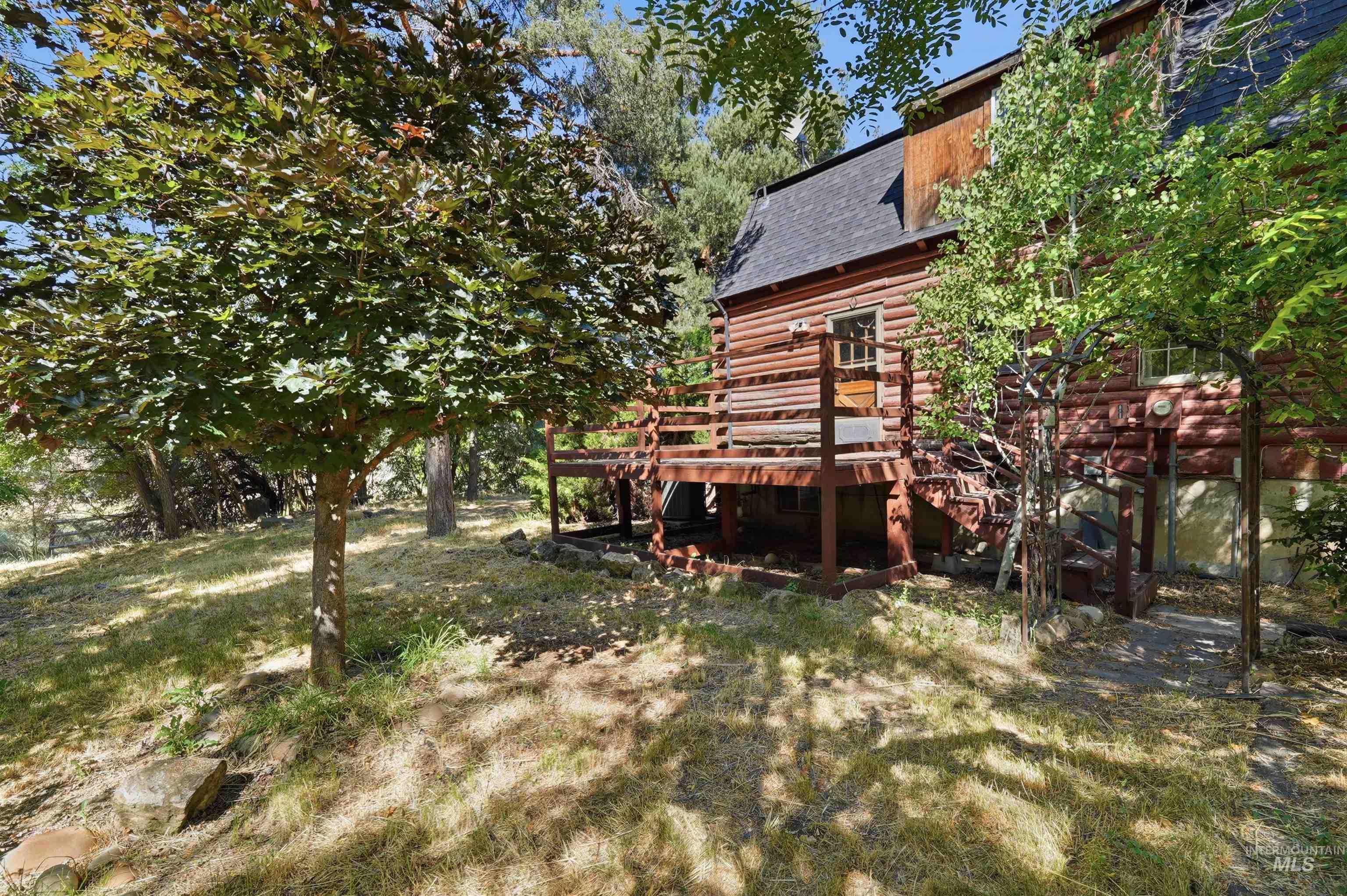 Log cabin exterior featuring a multi-level wood deck, dark shingle roof, and mature trees on a sloped lot