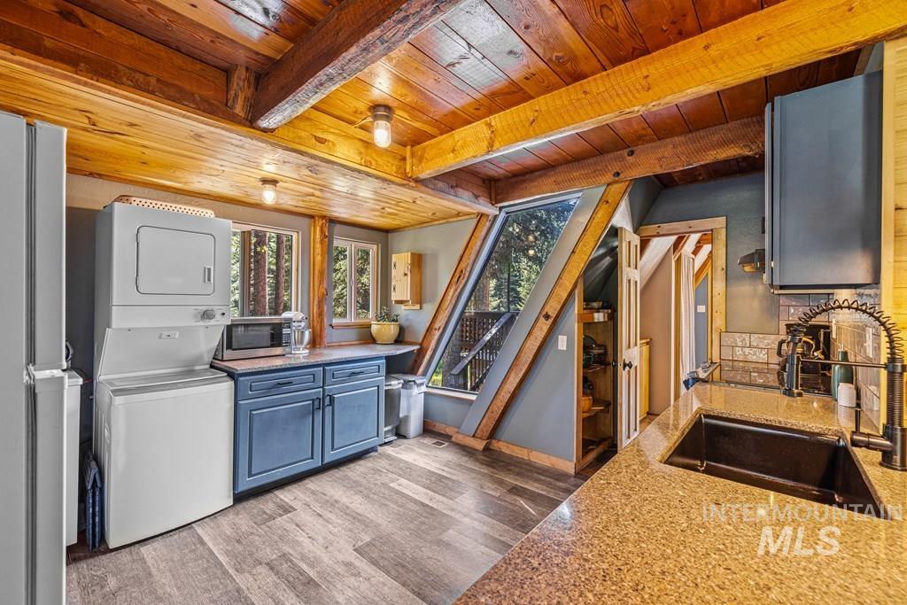 Kitchen featuring blue cabinets, light wood-type flooring, stacked washer and clothes dryer, appliances with stainless steel finishes, and a wooden ceiling with exposed beams