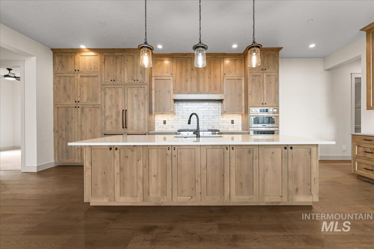 Kitchen with a large island, backsplash, decorative light fixtures, dark wood-type flooring, and light stone countertops