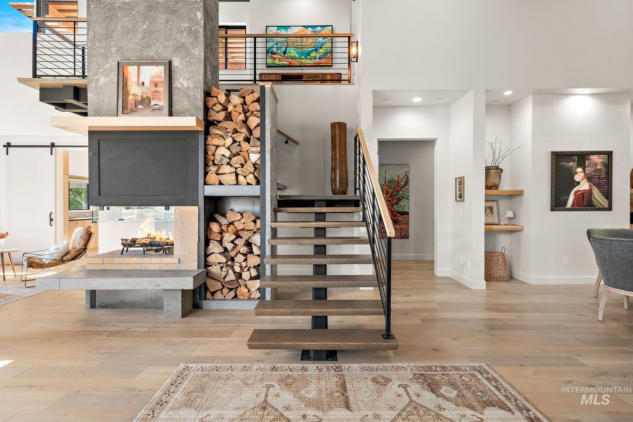 Open-concept living space showcasing a contemporary multi-level fireplace with integrated firewood storage, light wood-finish flooring, and a modern open-tread staircase with black iron railing