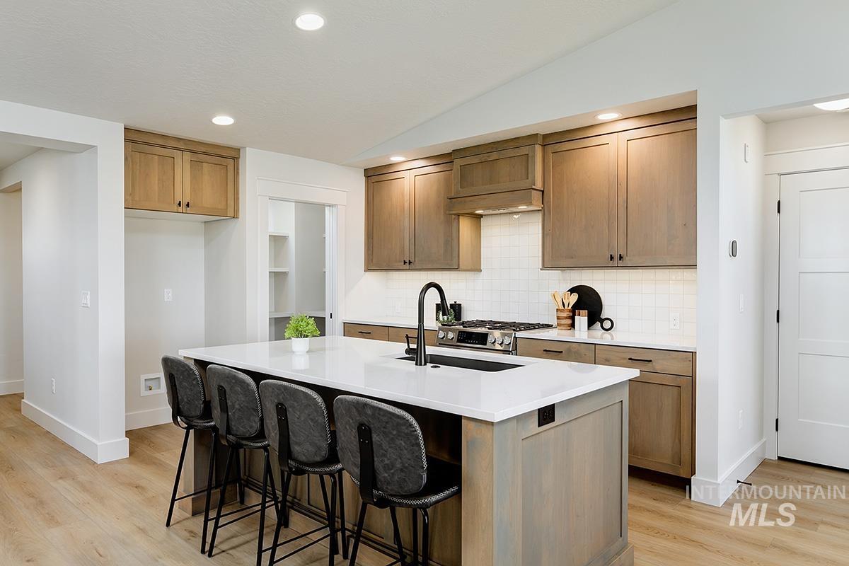 Kitchen featuring a large central island with a white countertop and built-in sink, natural wood-finish cabinetry, white subway tile backsplash, stainless steel gas range, and recessed lighting