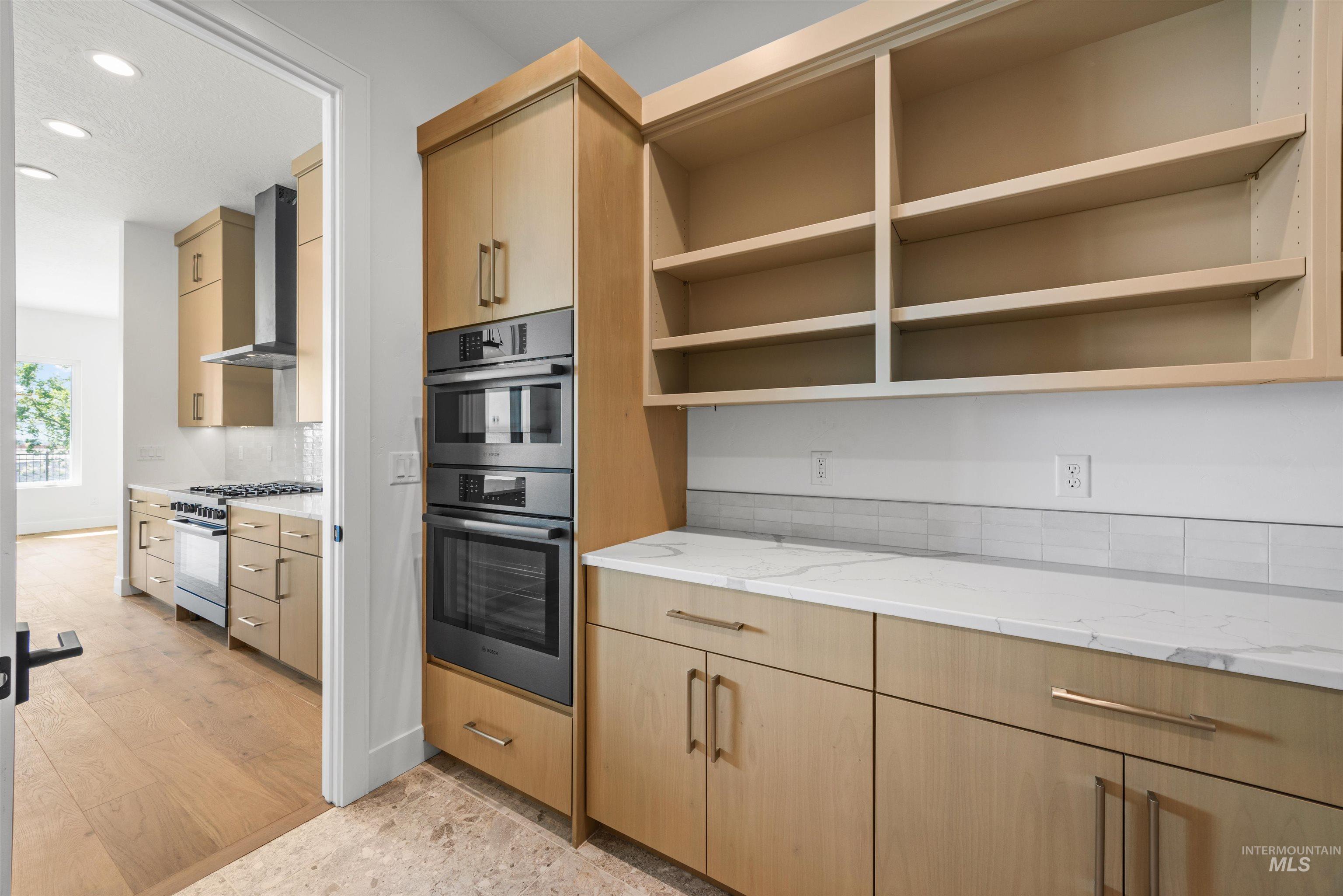 Kitchen featuring light stone countertops, light brown cabinetry, appliances with stainless steel finishes, wall chimney exhaust hood, and light wood finished floors