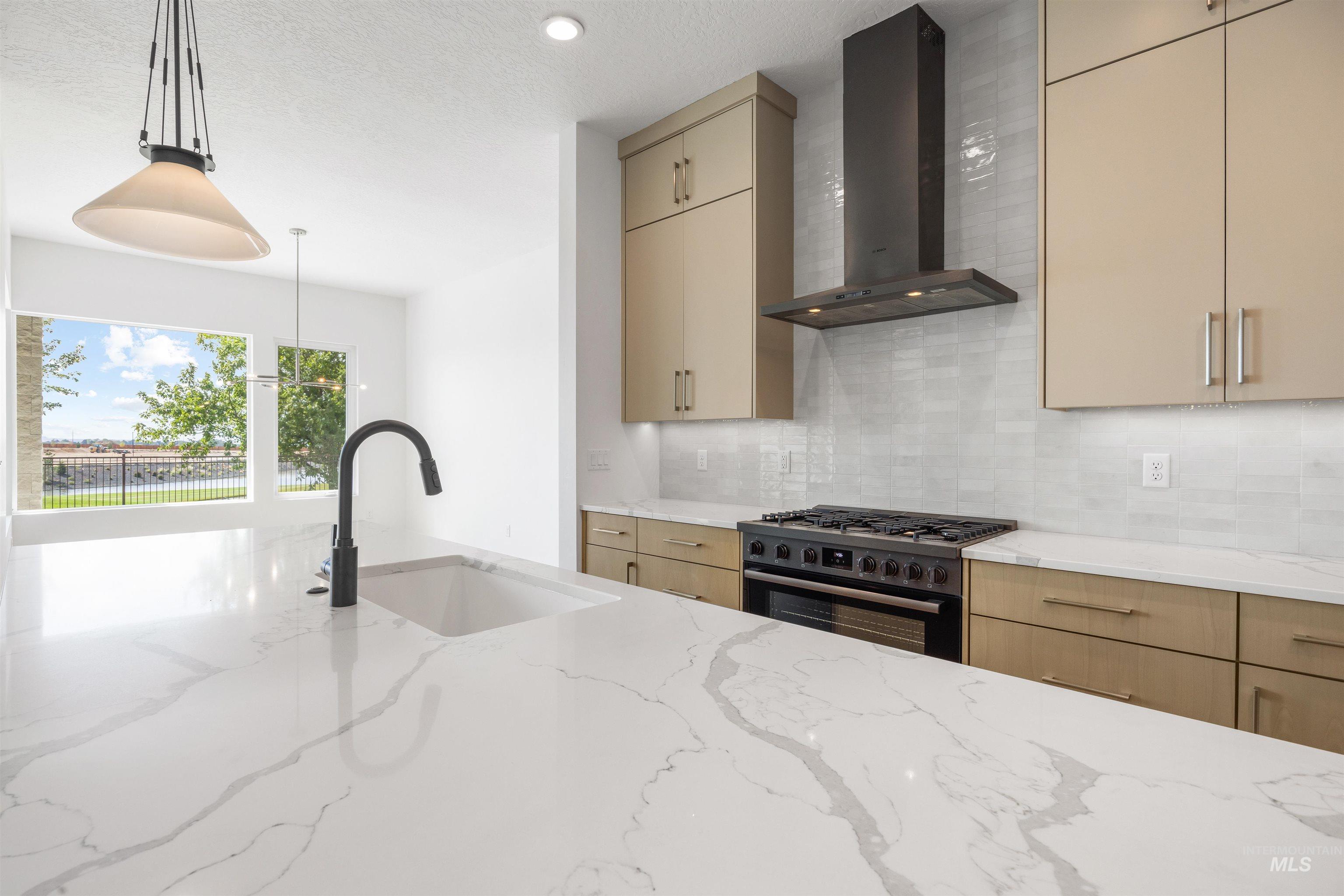 Kitchen featuring wall chimney range hood, black range with gas cooktop, light stone countertops, decorative backsplash, and hanging light fixtures