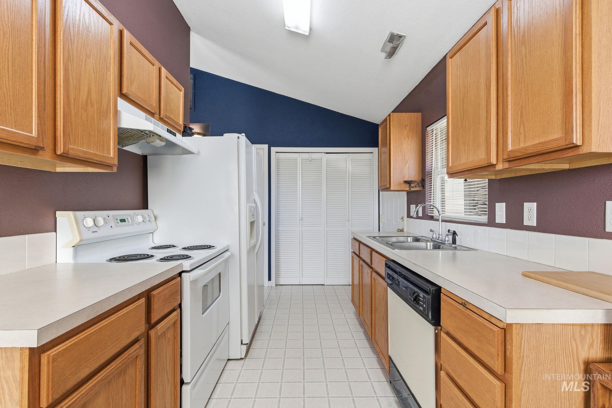 Kitchen with white appliances, lofted ceiling, under cabinet range hood, light tile patterned flooring, and light countertops
