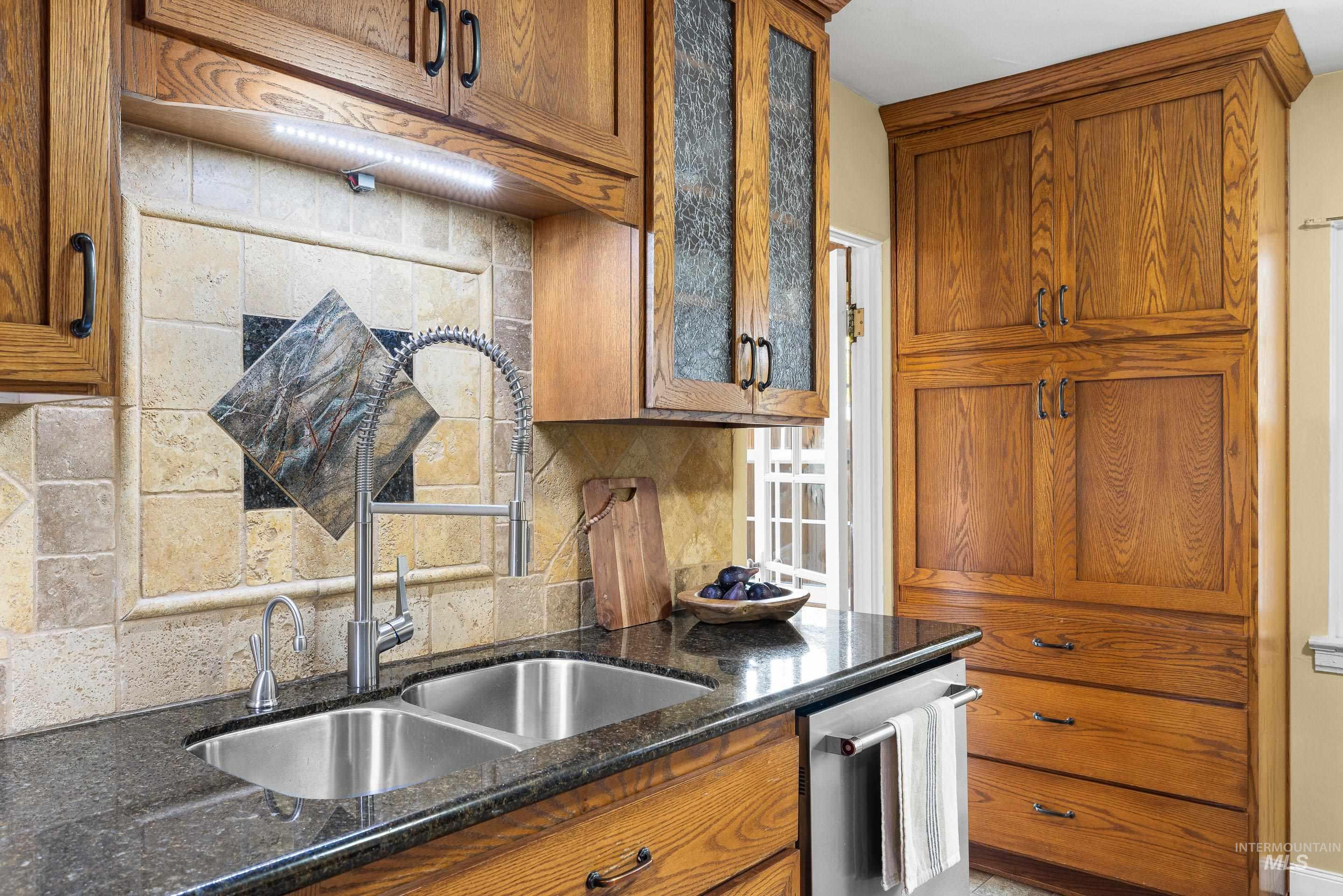 Kitchen with brown cabinetry, backsplash, dark stone counters, and dishwasher