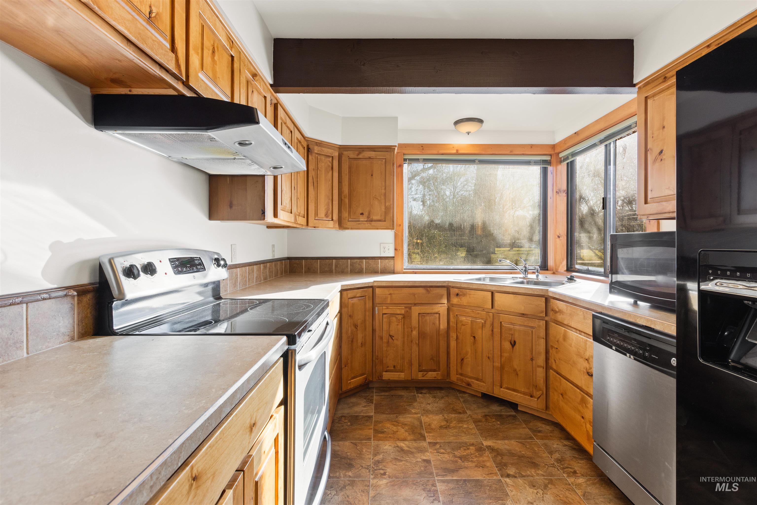 Kitchen with black appliances, under cabinet range hood, stone finish floors, light countertops, and brown cabinets