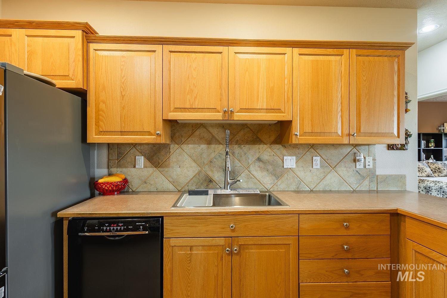 Kitchen with black dishwasher, light countertops, freestanding refrigerator, decorative backsplash, and brown cabinetry