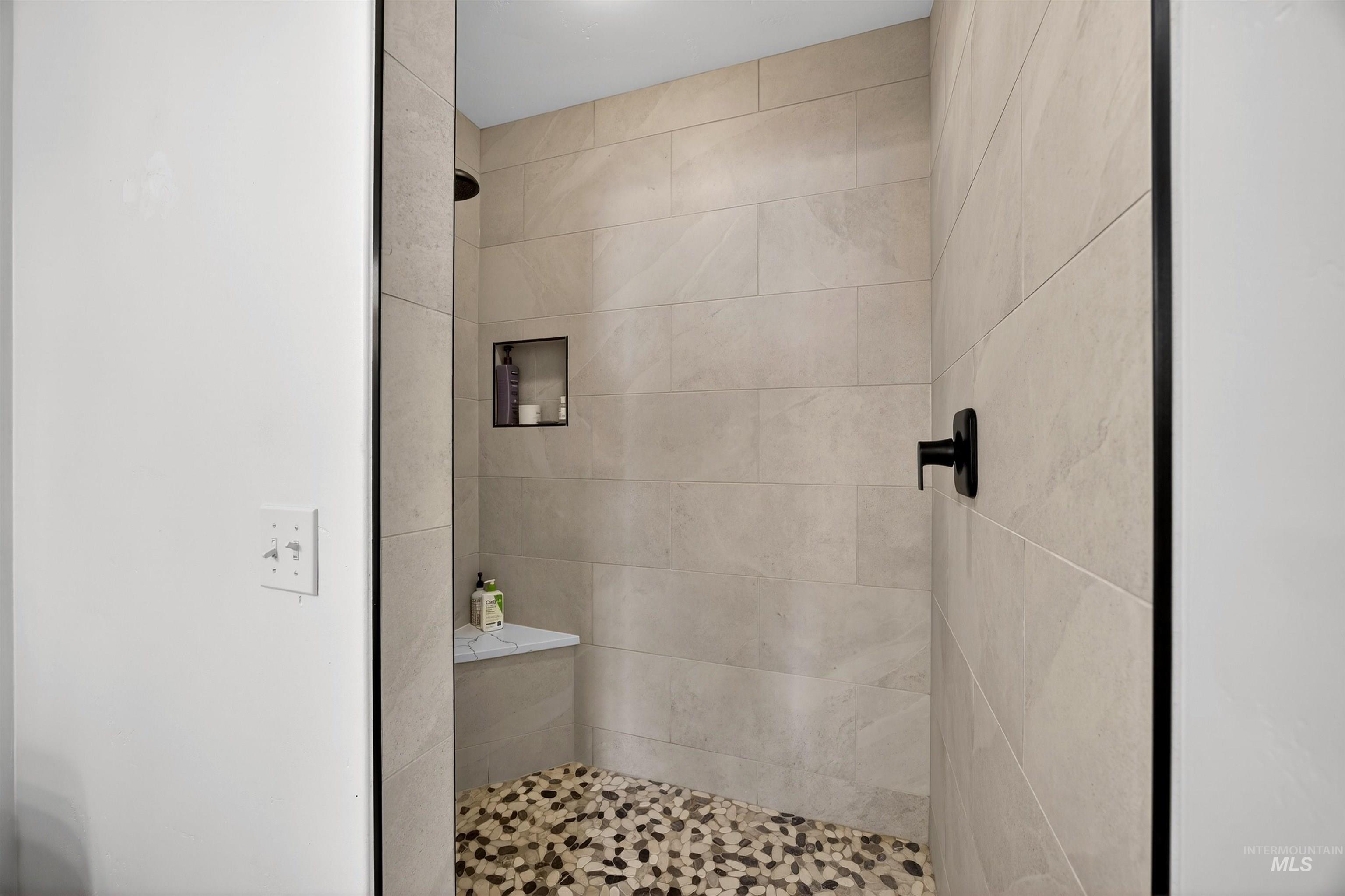 Walk-in shower featuring large format tile walls, a pebble floor, built-in shelving, a corner bench, and matte black fixtures