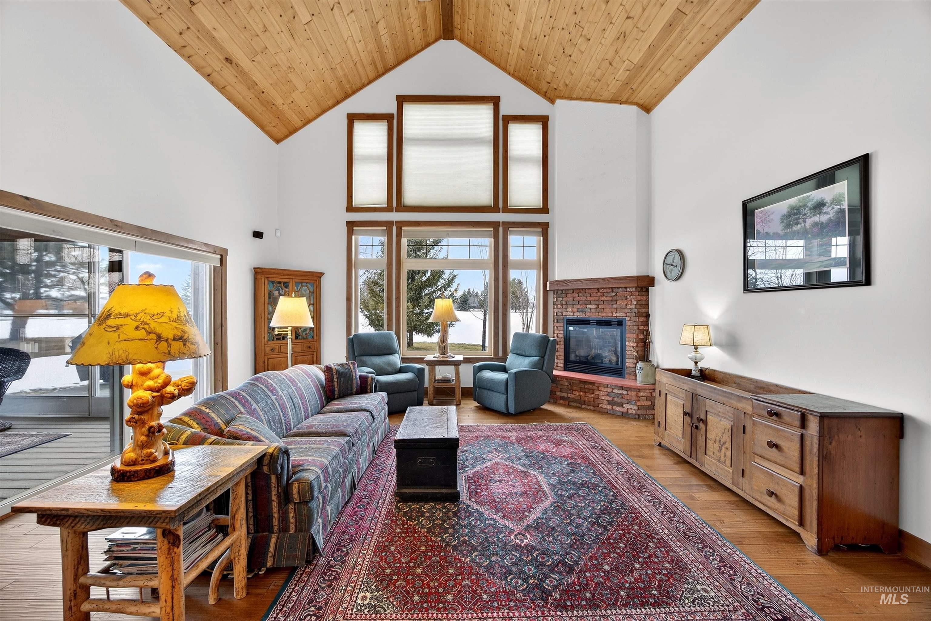 Spacious living area featuring a vaulted wood-paneled ceiling, large picture windows, a brick-faced fireplace, and hardwood flooring