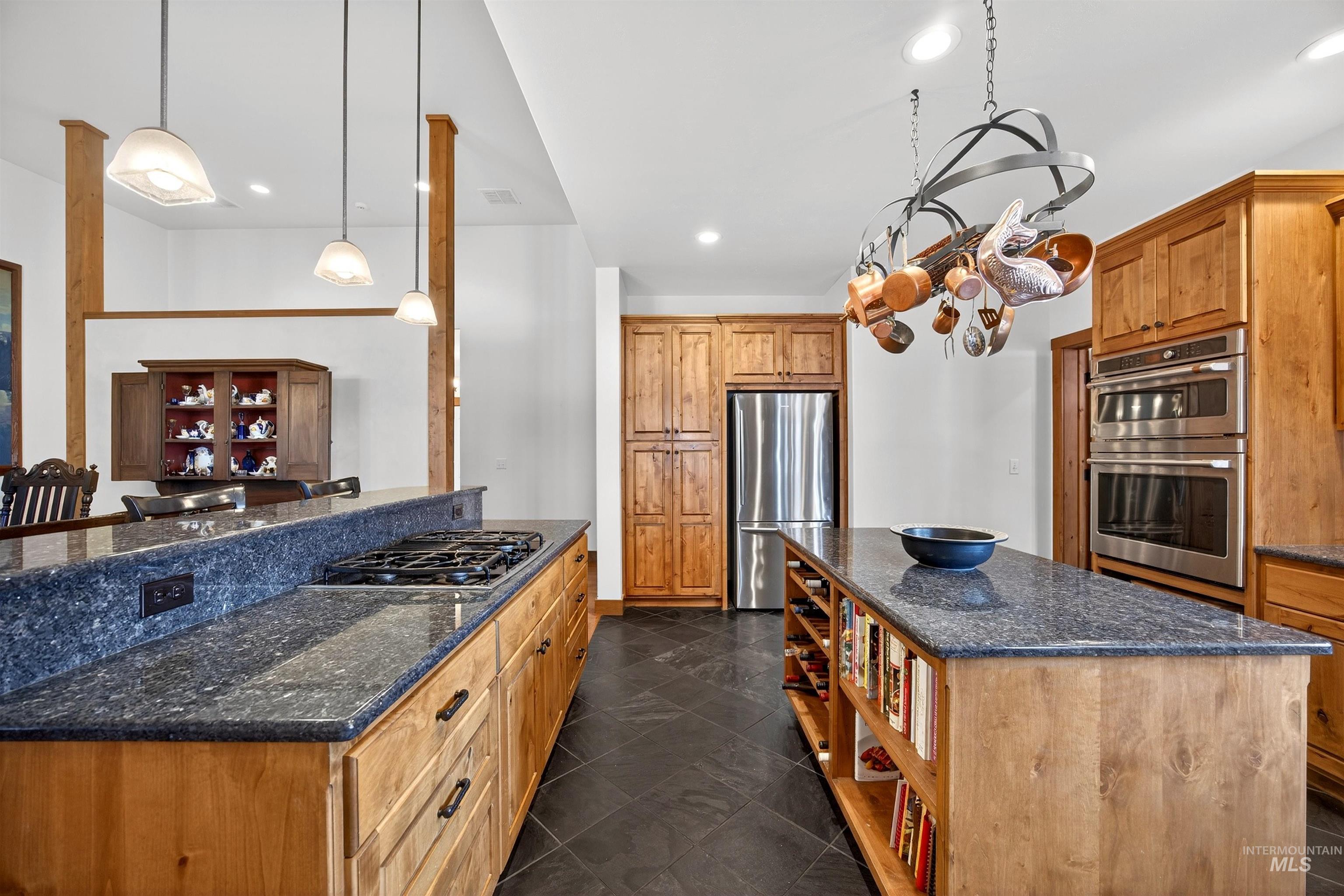 Kitchen featuring an island with integrated shelving, dark stone countertops, wood-finish cabinetry, stainless steel appliances, and dark tile flooring